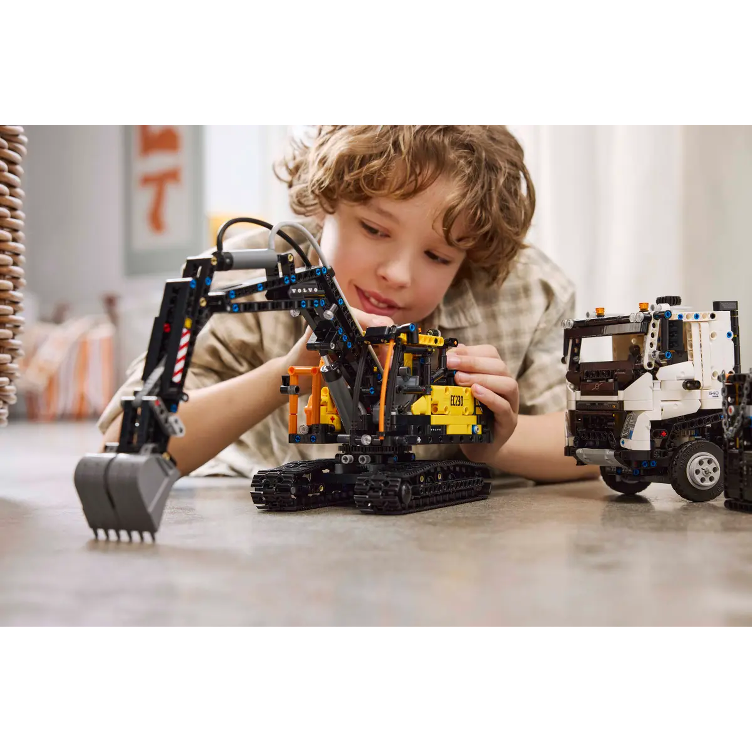 A boy with curly hair is playing with toy construction vehicles. The toys include an excavator and a truck, on a table.