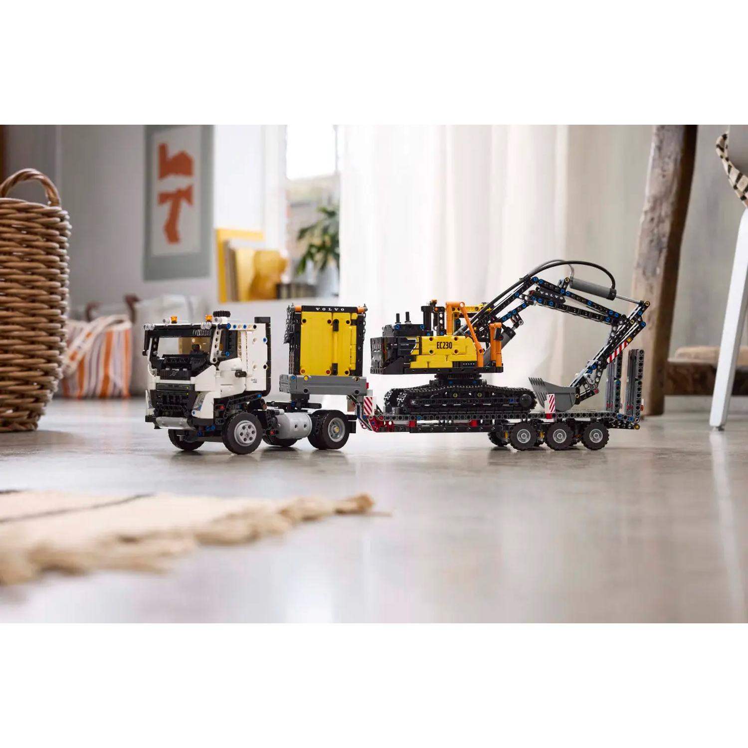 A Lego Technic truck pulling an excavator on a trailer is on a floor. A basket and a painting are in the background.