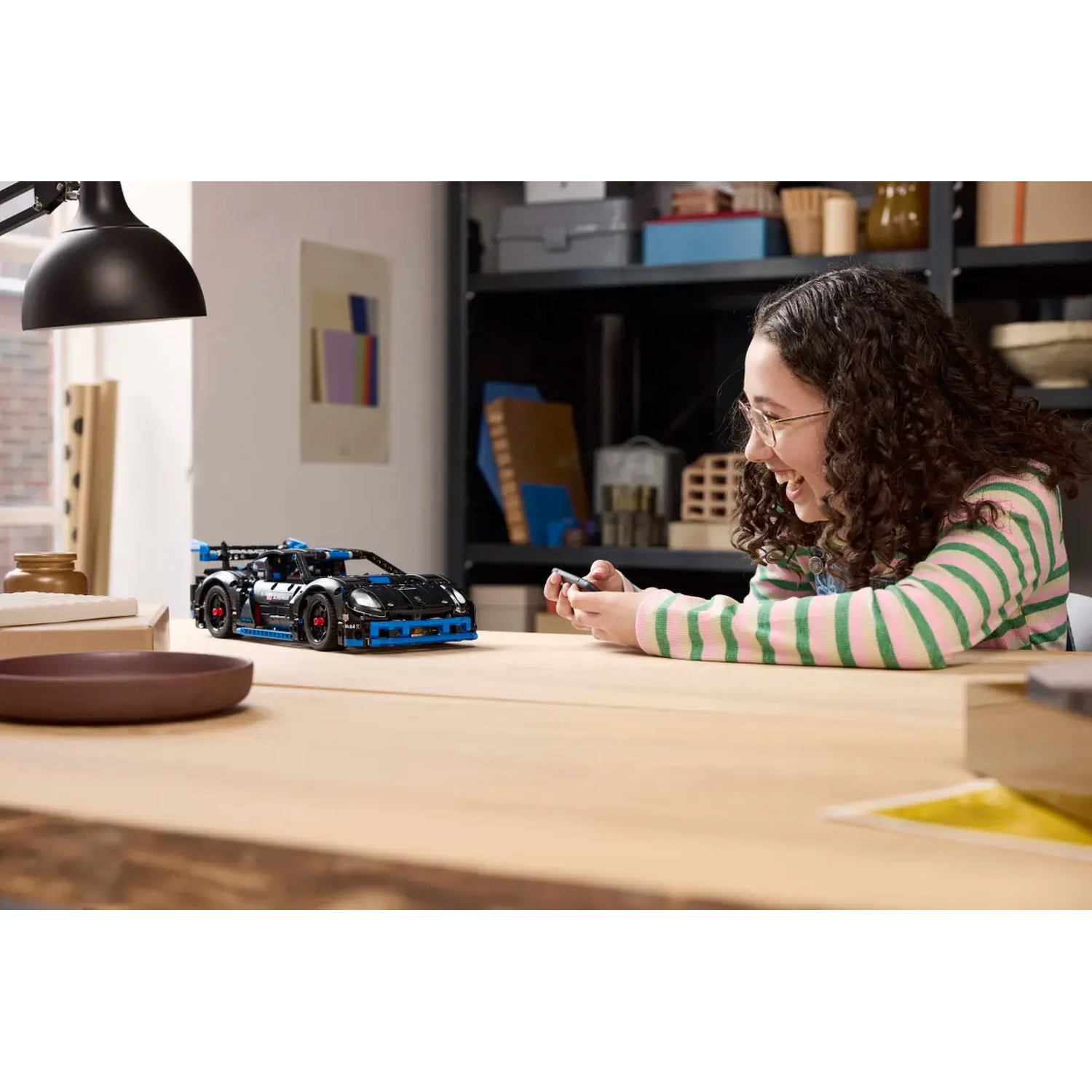 A girl smiles while operating a remote. A black Lego car sits on a wooden table. A lamp is to the left.