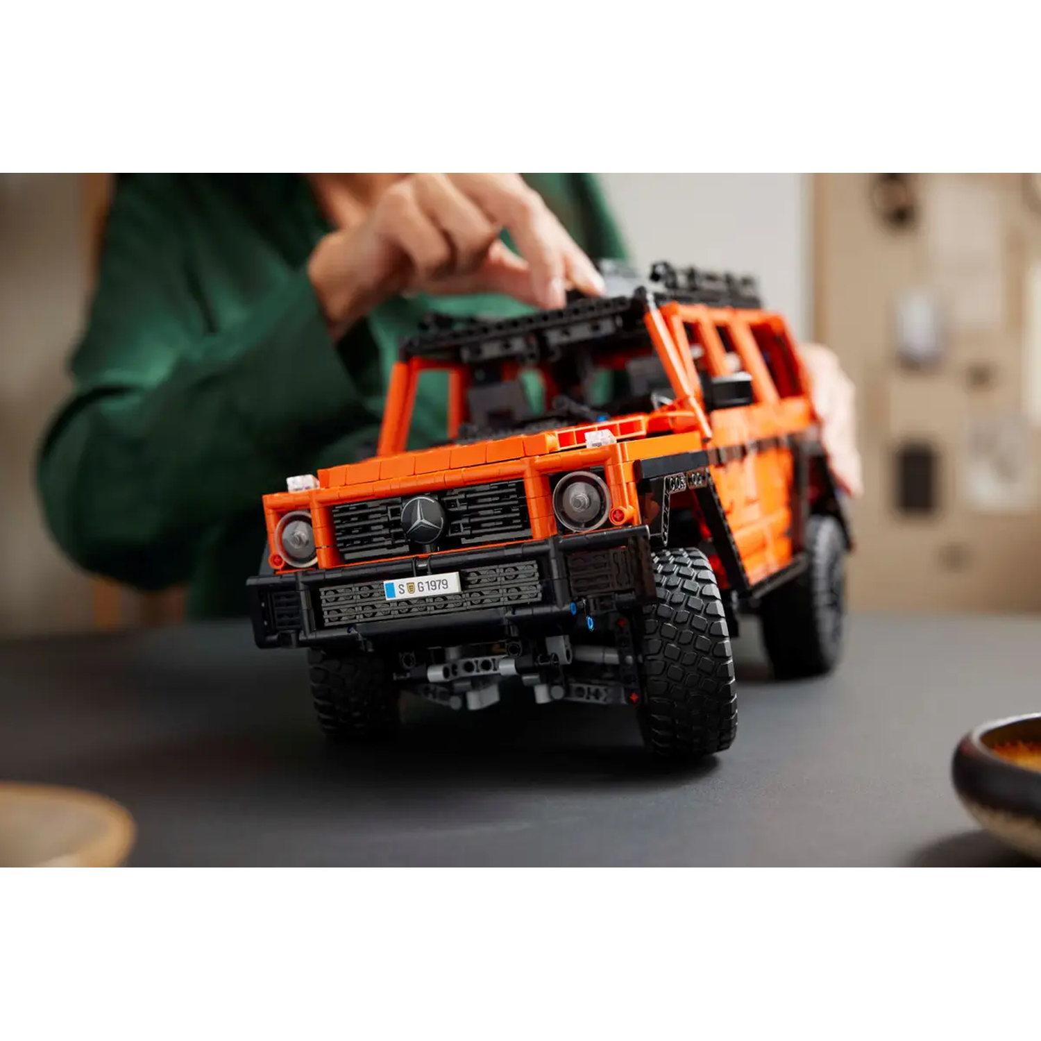 An orange Lego model of a Mercedes-Benz G-Wagon. A hand is touching the top of the car, which is on a dark surface.