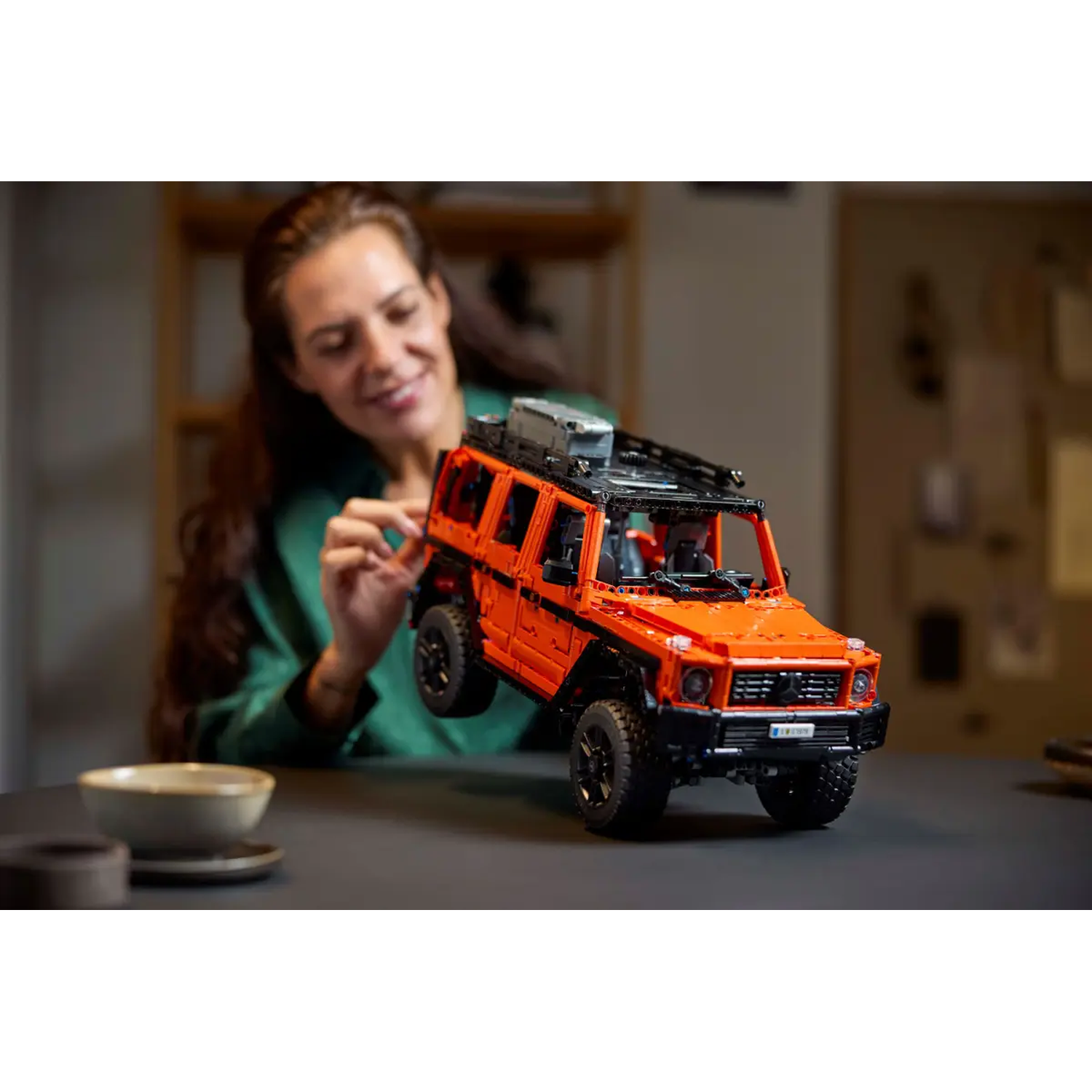 A woman holds an orange toy SUV. A table has a bowl and a coaster. The background is slightly blurred with warm light.