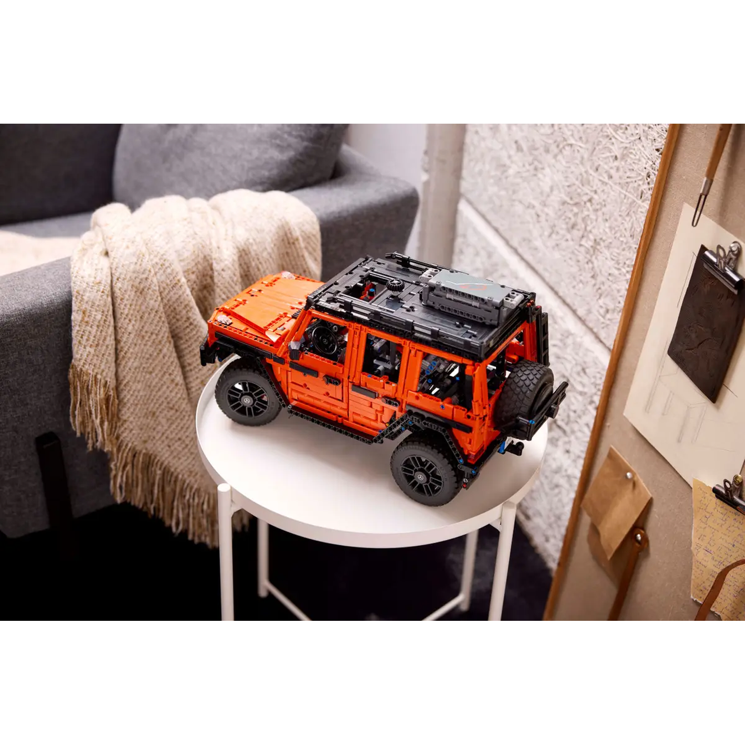 An orange Lego model SUV sits on a white table. A gray sofa and a bulletin board are in the background.