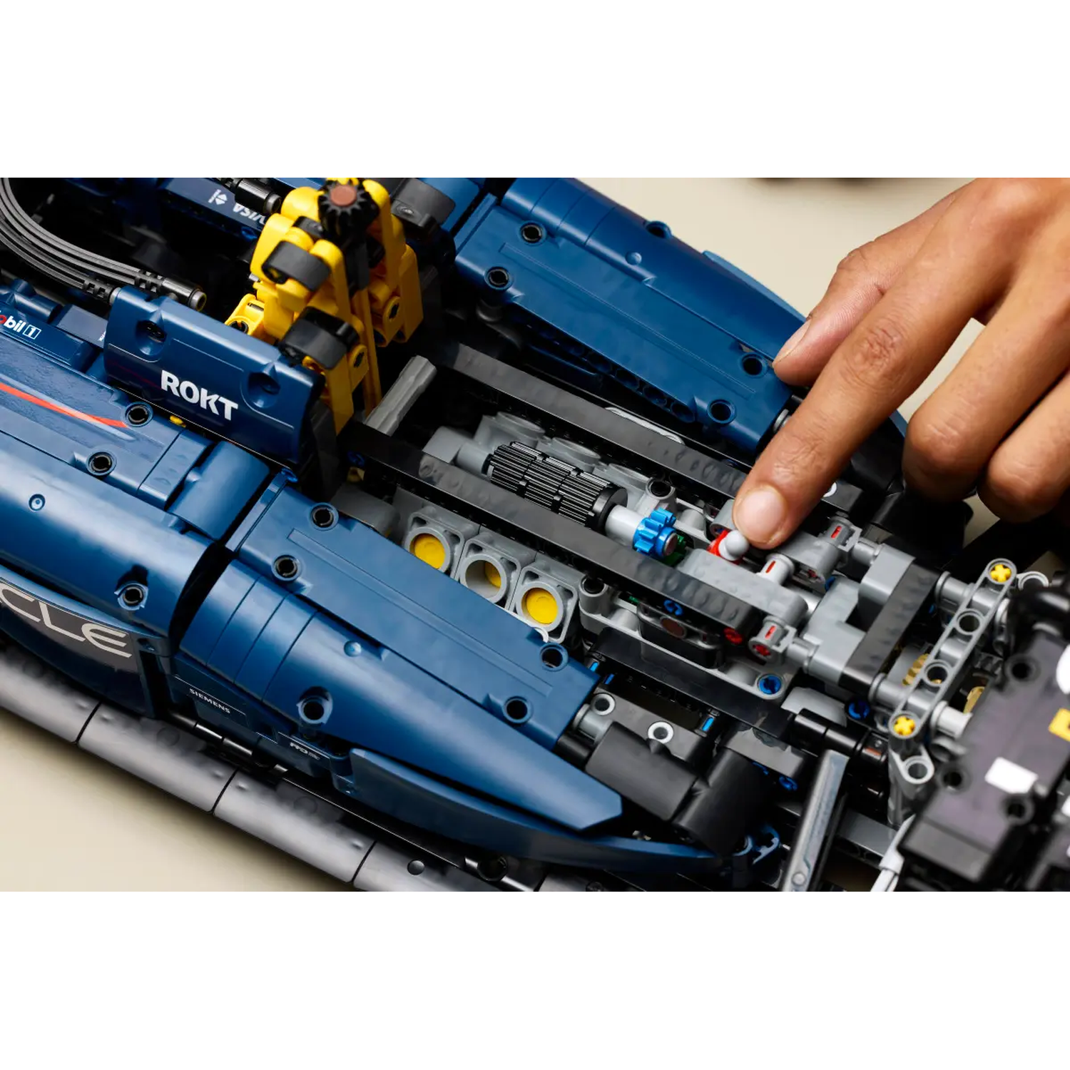 Een hand wijst naar de motor van een blauwe Lego-auto. Een gele Lego-figuur zit erachter. De auto heeft opschriften.
