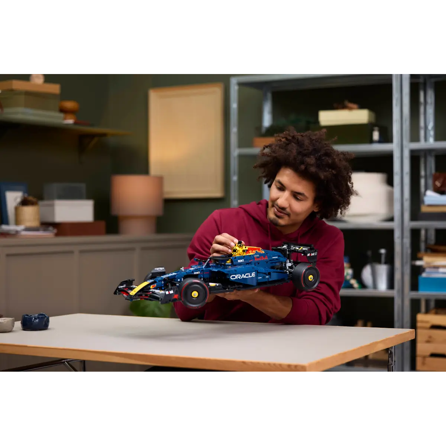 Een persoon houdt een blauw en geel raceauto-model vast. Ze bevinden zich in een kamer met planken en een tafel.