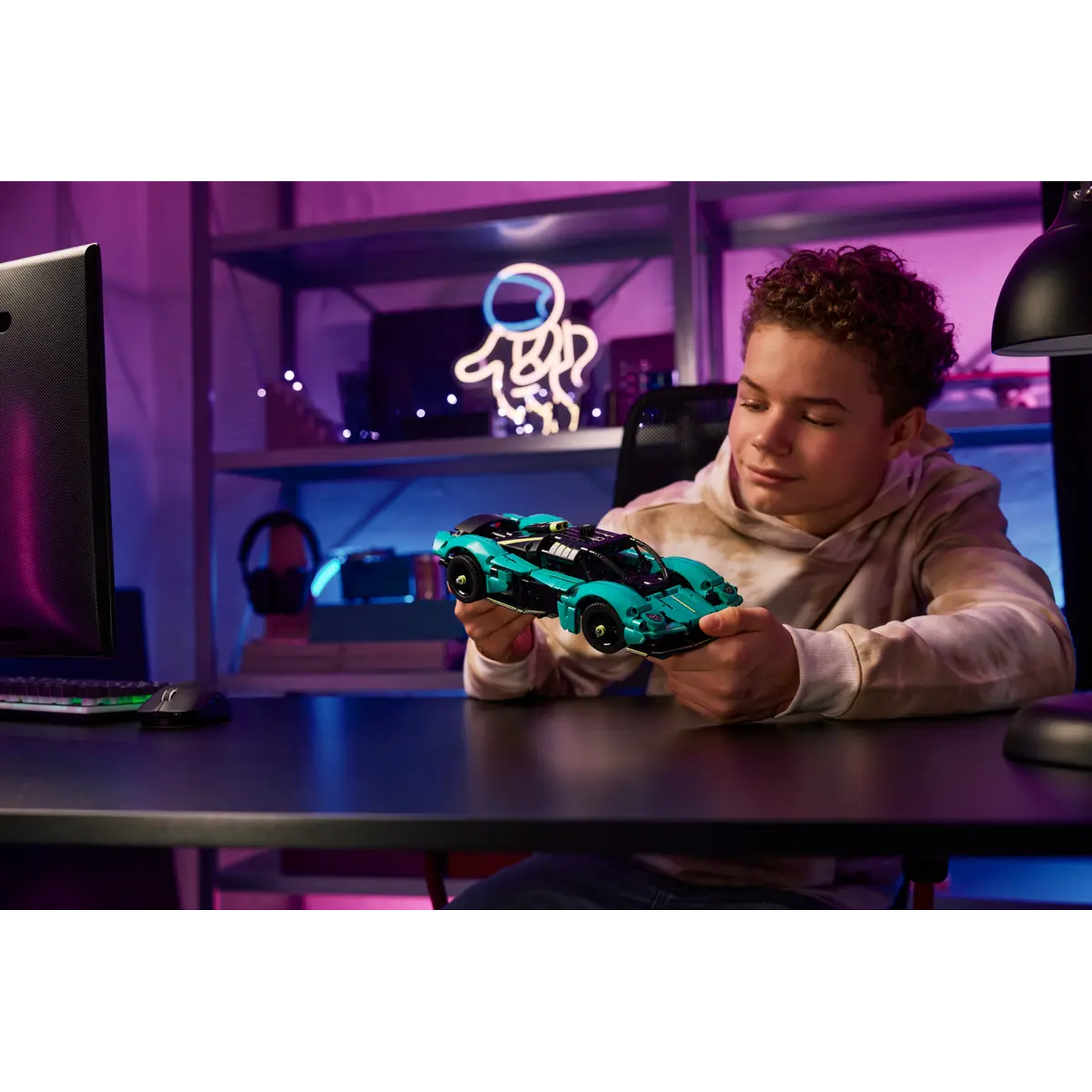 A boy holds a teal Lego car. A computer and desk lamp are on the desk. Neon astronaut art decorates the background.