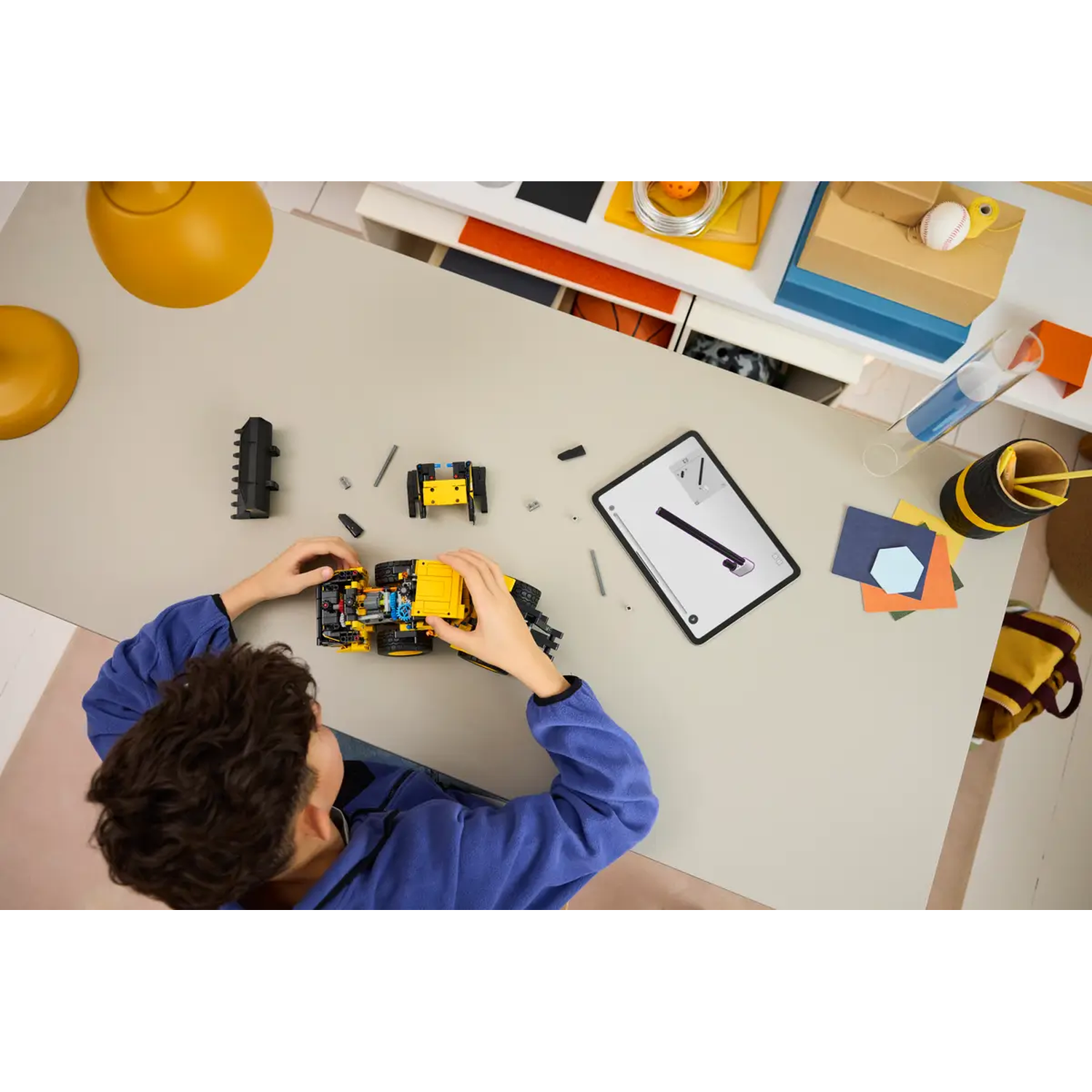 A child assembles a yellow and black toy vehicle on a light gray desk. Various objects are scattered around.