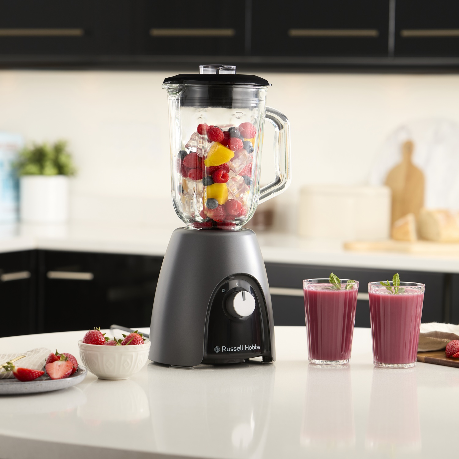 Un frullatore con frutta e ghiaccio, accanto a due frullati e fragole su un tavolo in cucina.