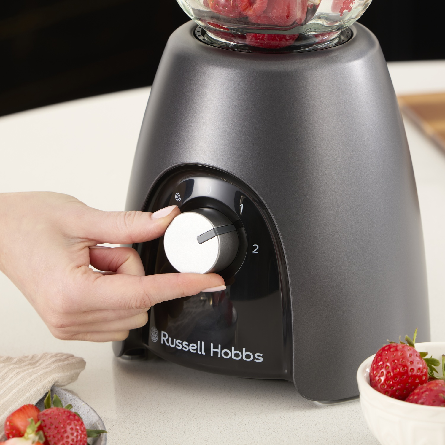 Una mano gira il quadrante di un frullatore Russell Hobbs. Il frullatore contiene fragole, e ci sono più fragole in una ciotola.