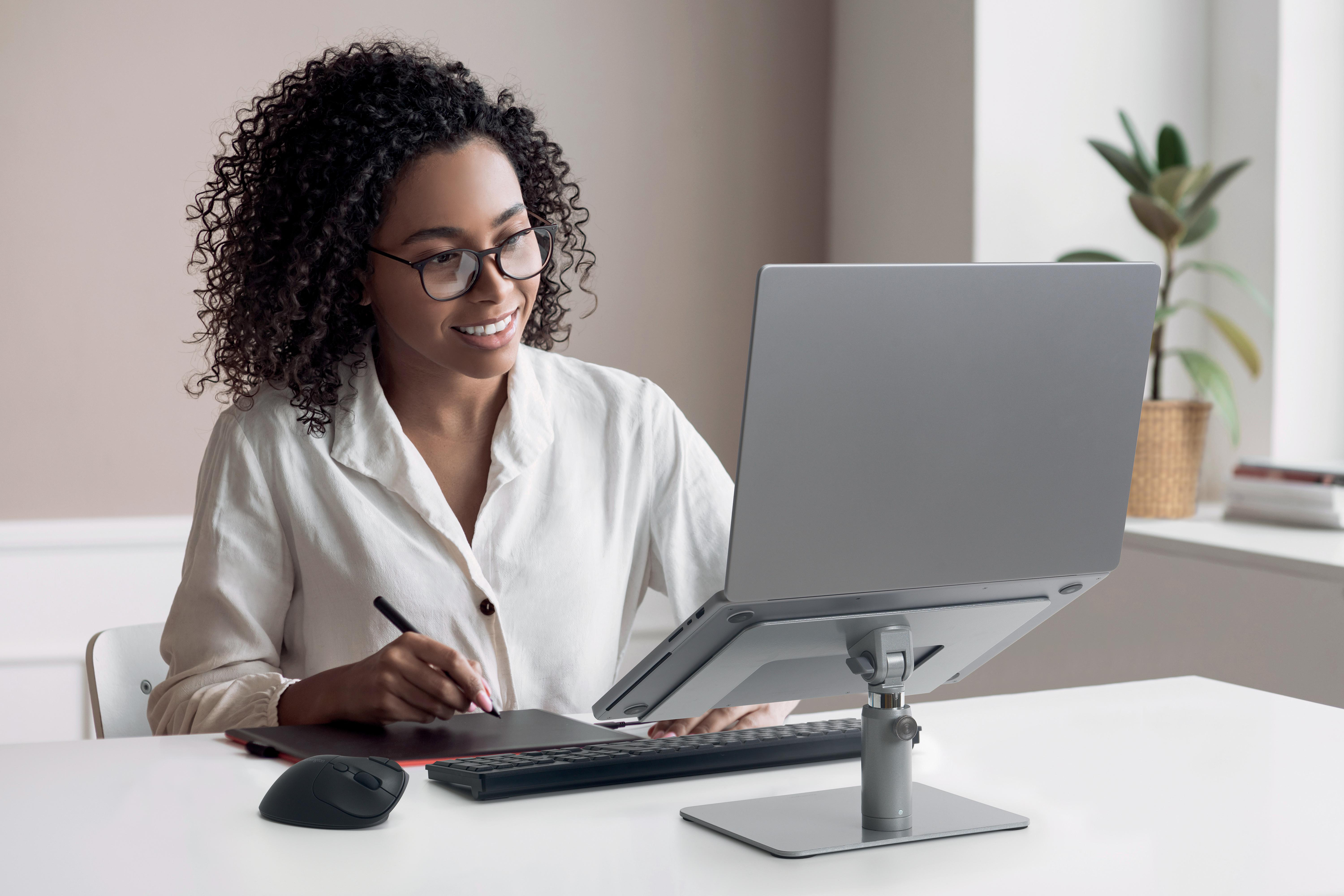 Frau mit Brille lächelt bei der Arbeit am Laptop. Sie benutzt einen Stift auf einem Tablet, mit einer Maus auf dem Tisch.