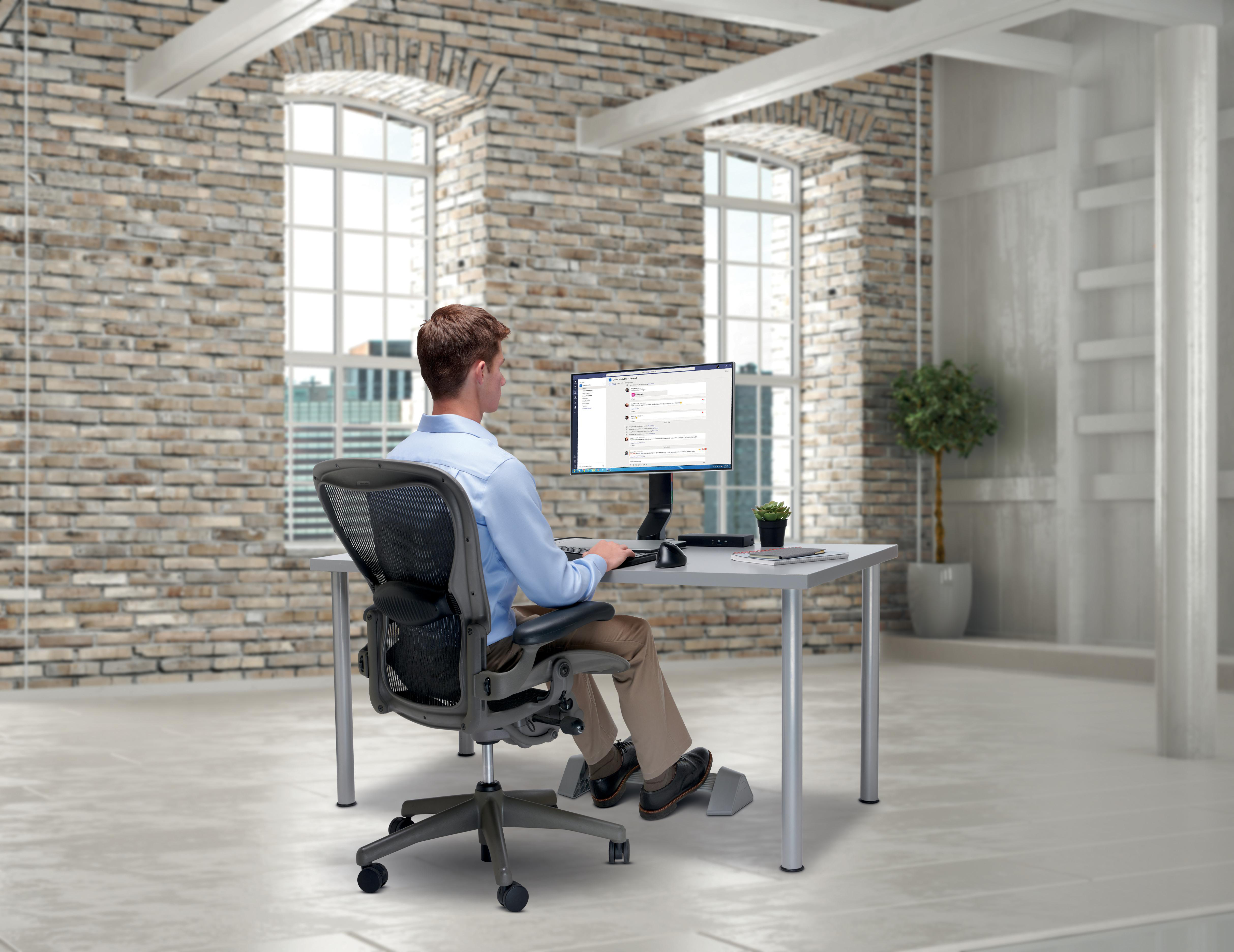 Ein Mann sitzt an einem Schreibtisch und blickt auf einen Computer. Hintergrund: Backsteinmauer, Fenster. Schreibtisch mit Monitor, Maus und Pflanze.