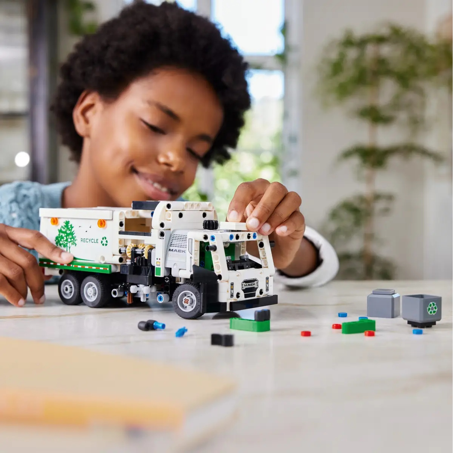 A smiling child is building a toy truck. The truck is white, green, and black. Various Lego pieces are on the table.