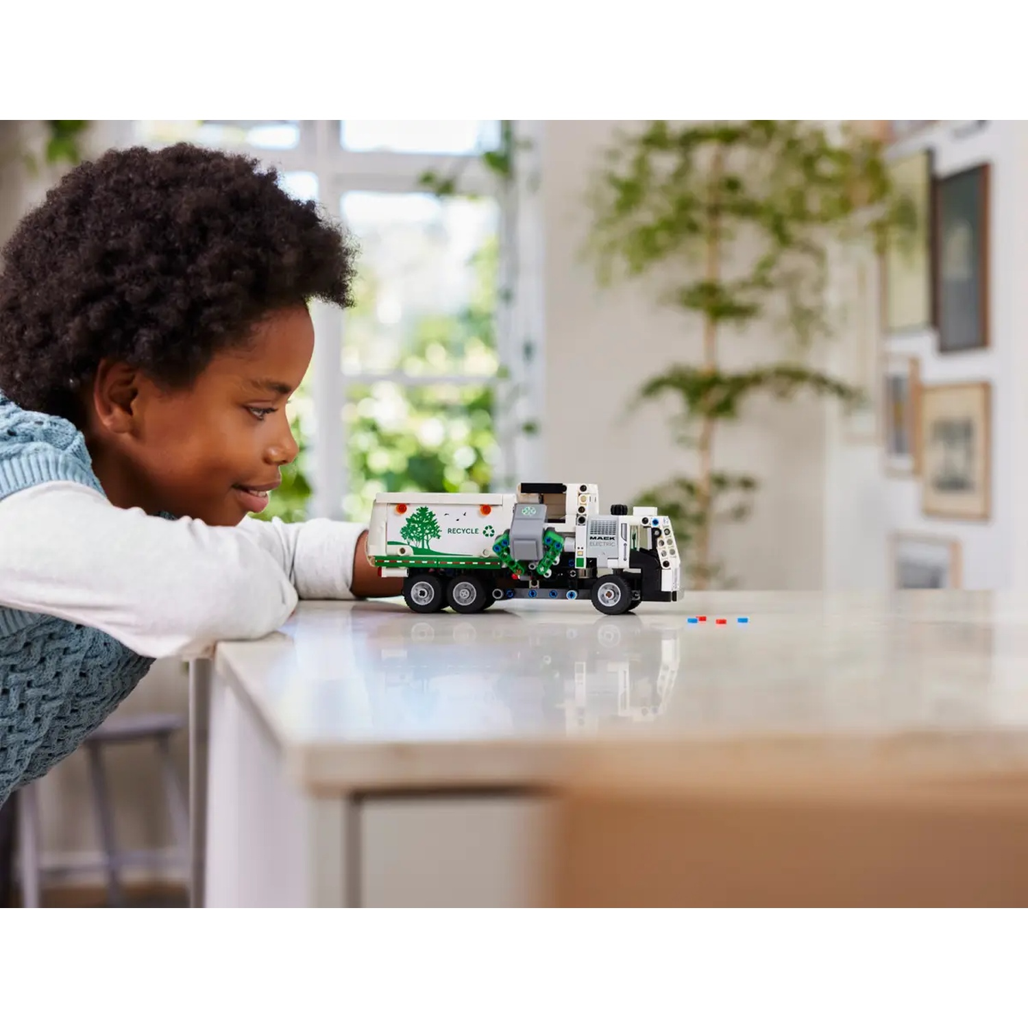 A child looks at a toy garbage truck on a counter. The truck is white, with green accents, and the word recycle on it.