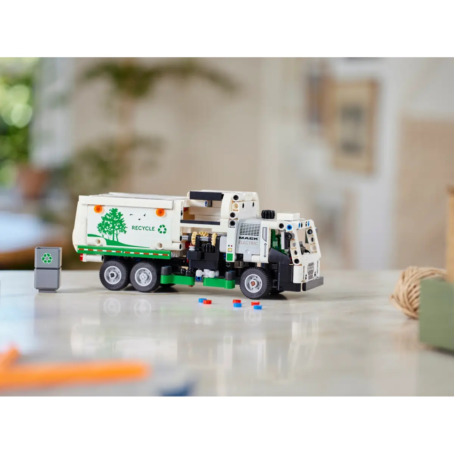 A toy garbage truck with green and white colors, recycle logo, and a small bin on the left. The background is blurred.