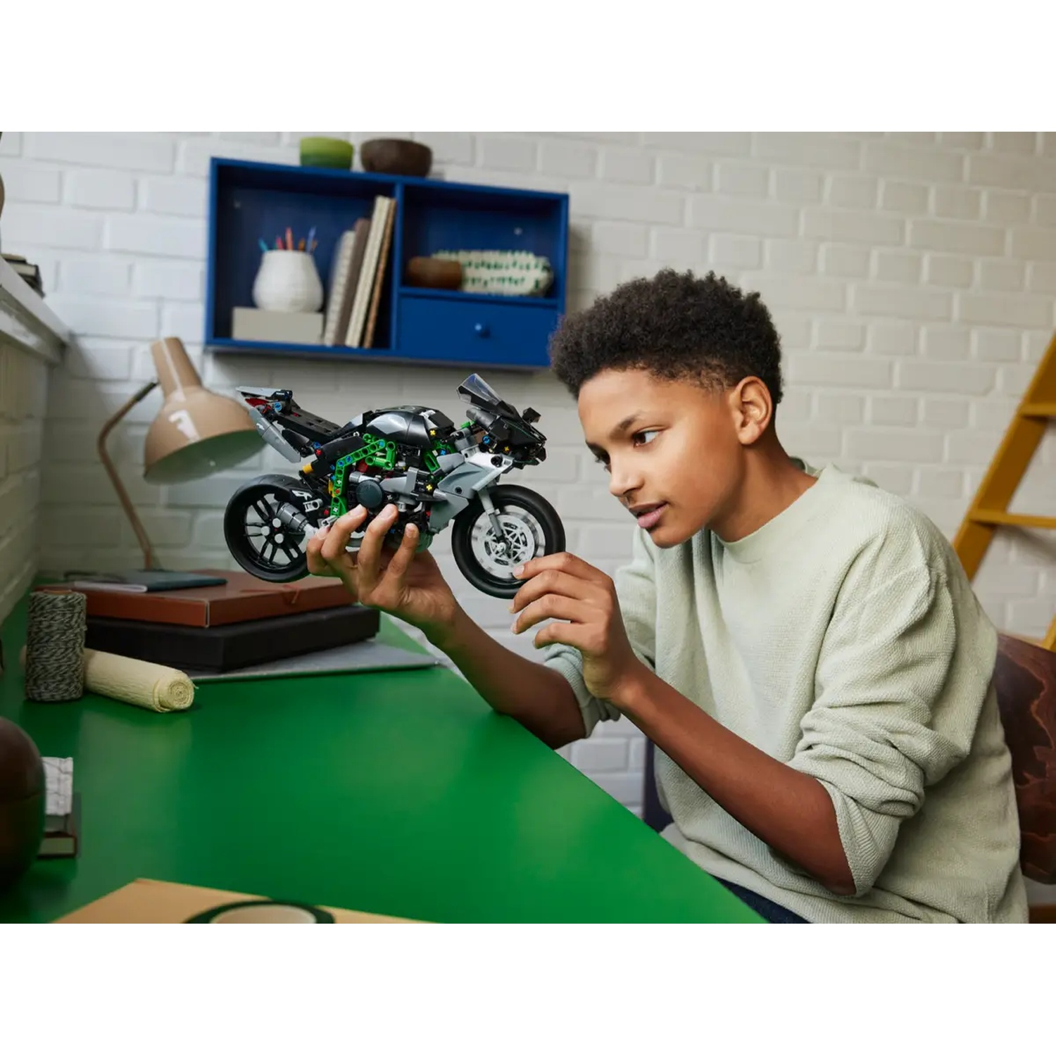 Een tiener bekijkt een motorfietsmodel op een groen bureau. Achtergrond omvat een blauwe plank, lamp en boeken.
