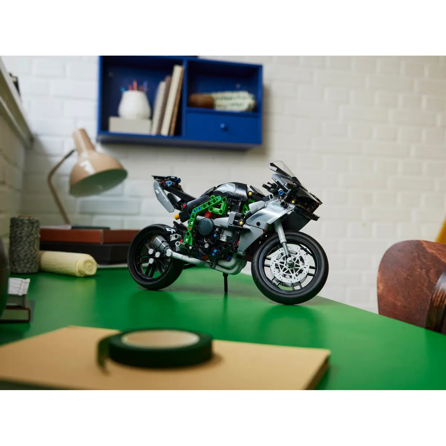 Een Lego-motorfiets staat op een groene tafel. Een rol plakband en een notitieblok staan op de voorgrond. Een blauwe plank en een lamp op de achtergrond.
