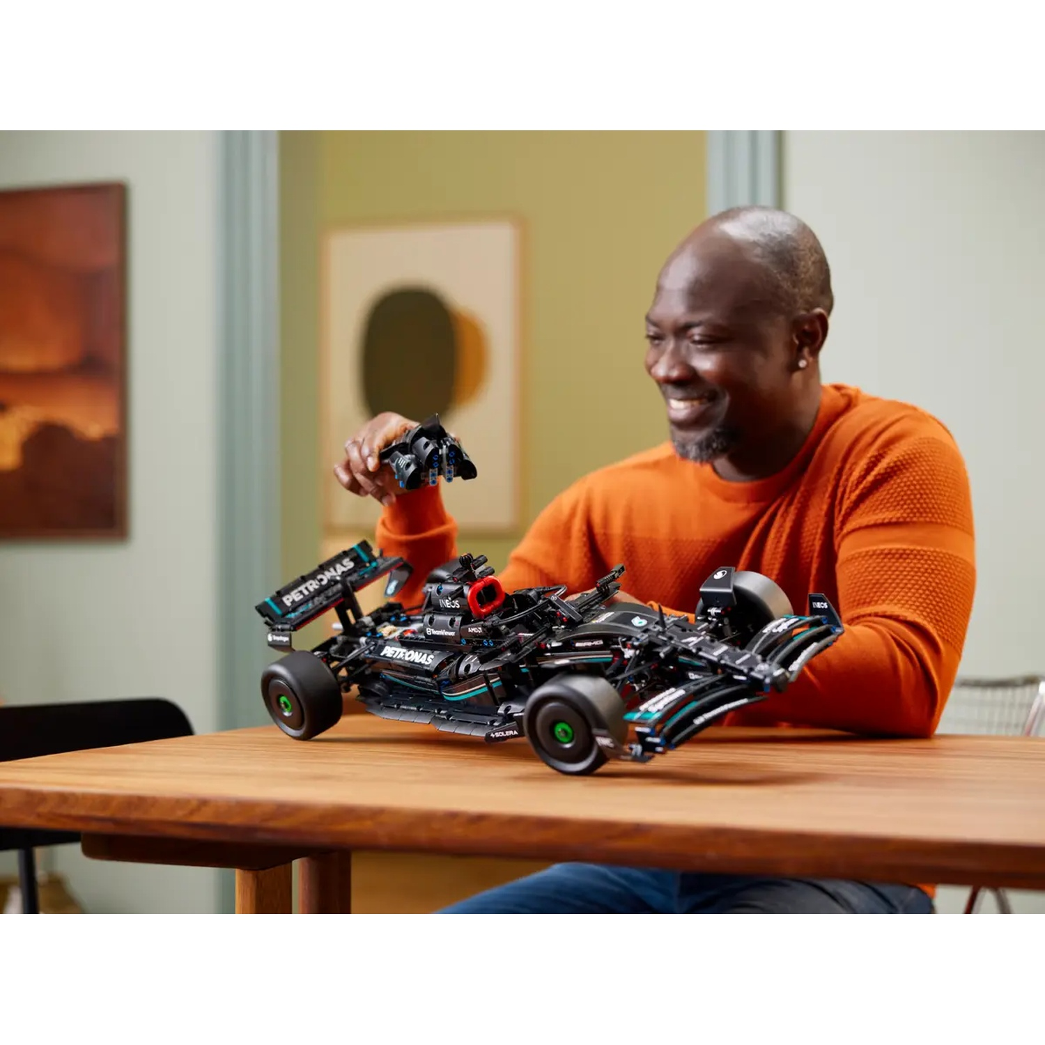 Un homme sourit en tenant une maquette de voiture de course noire et sarcelle. La voiture est sur une table en bois. Le fond comprend de l'art.