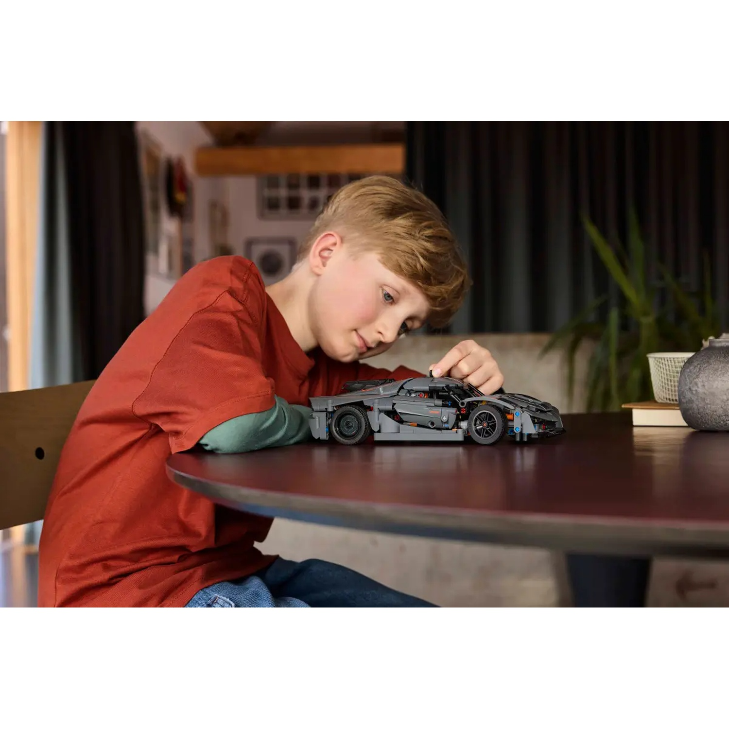 A boy in a red shirt is looking at a gray toy car on a round brown table. Background includes a plant and picture frames.