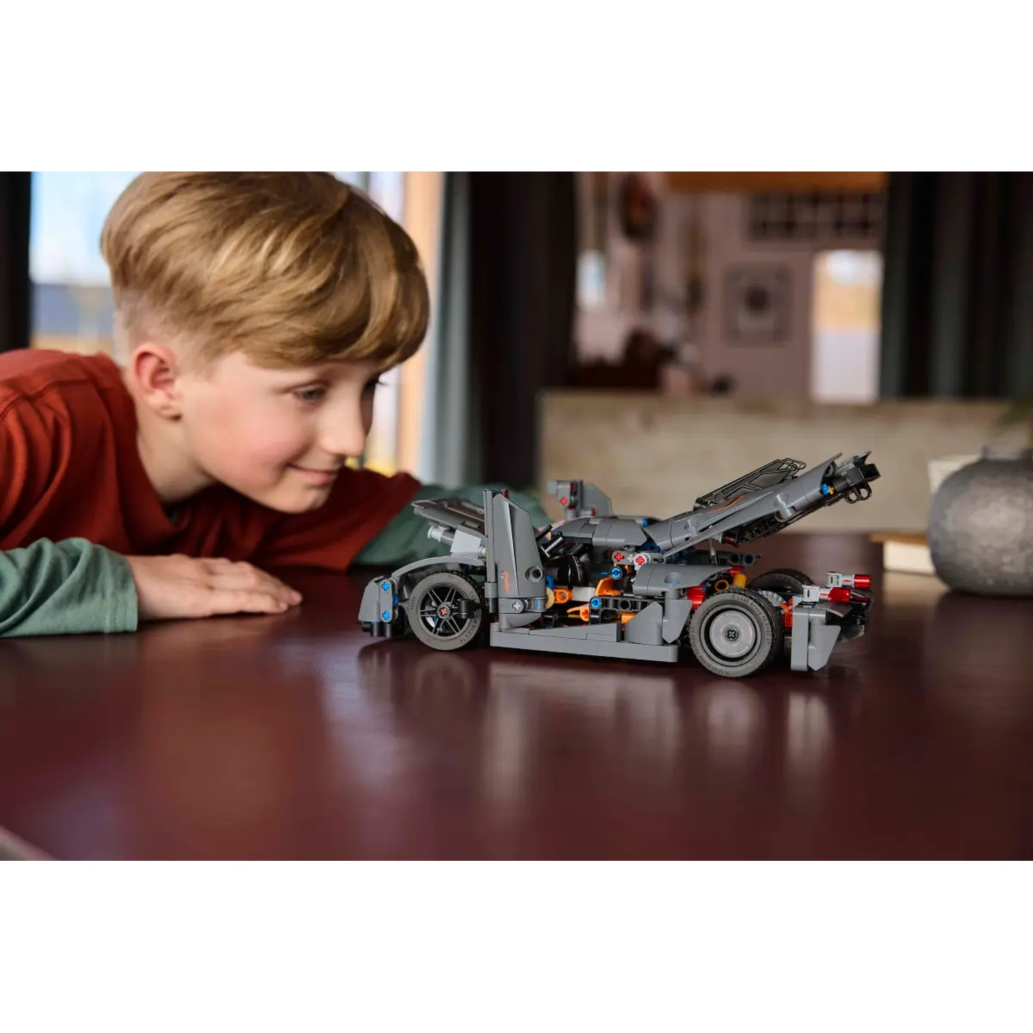 A boy looks at a grey LEGO car on a table. The car has black wheels and orange details. The background is blurred.