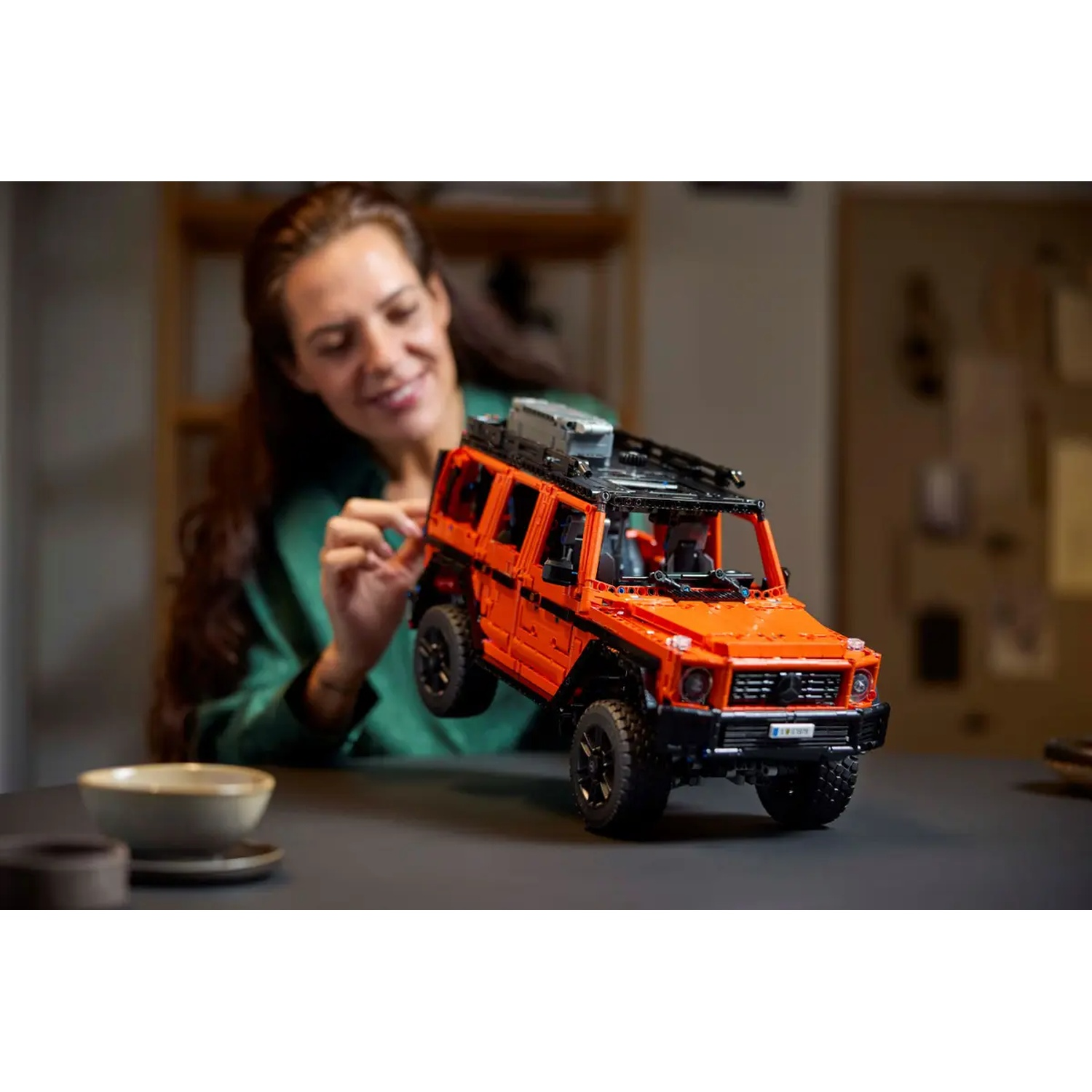 Une femme sourit en tenant une voiture jouet orange. La voiture est sur une table avec un bol et un dessous de verre. L'arrière-plan est flou.