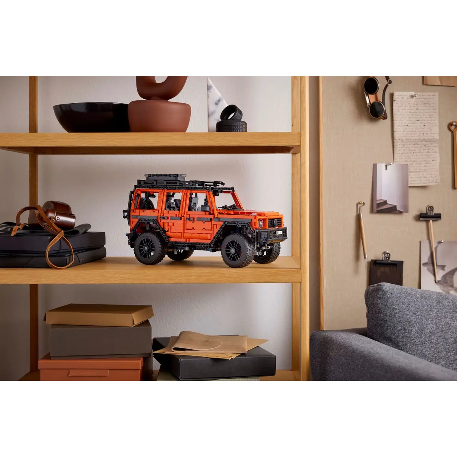Un Jeep Lego orange est posé sur une étagère. D'autres objets sur l'étagère sont un bol noir, des boîtes et un sac d'appareil photo. Un canapé gris est visible.