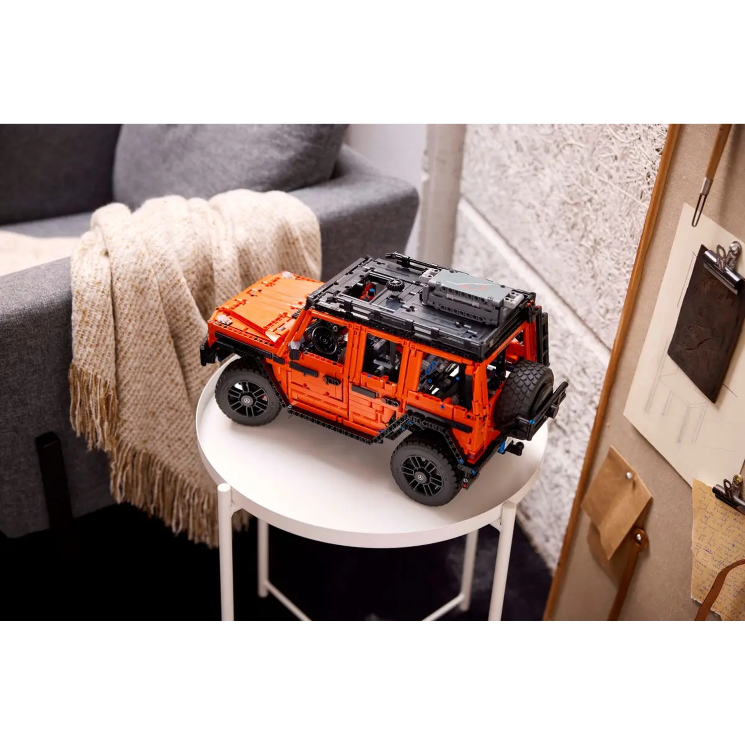 Un modèle de SUV Lego orange sur une table blanche. Un canapé gris, une couverture et un tableau d'affichage sont en arrière-plan.