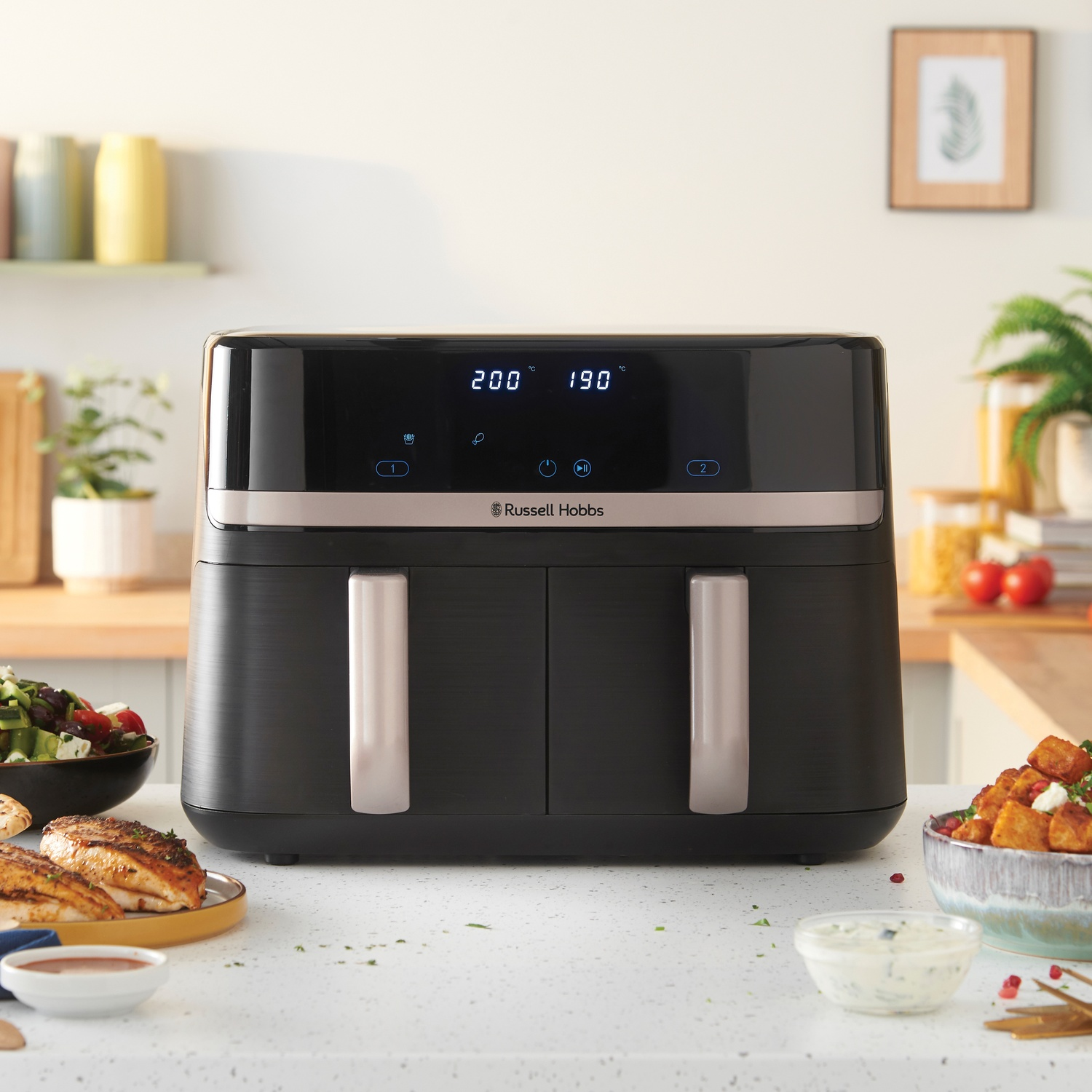 Friggitrice ad aria nera con display digitale. Cibo, tra cui pollo e insalata, sul bancone. Sfondo cucina con piante e decorazioni.