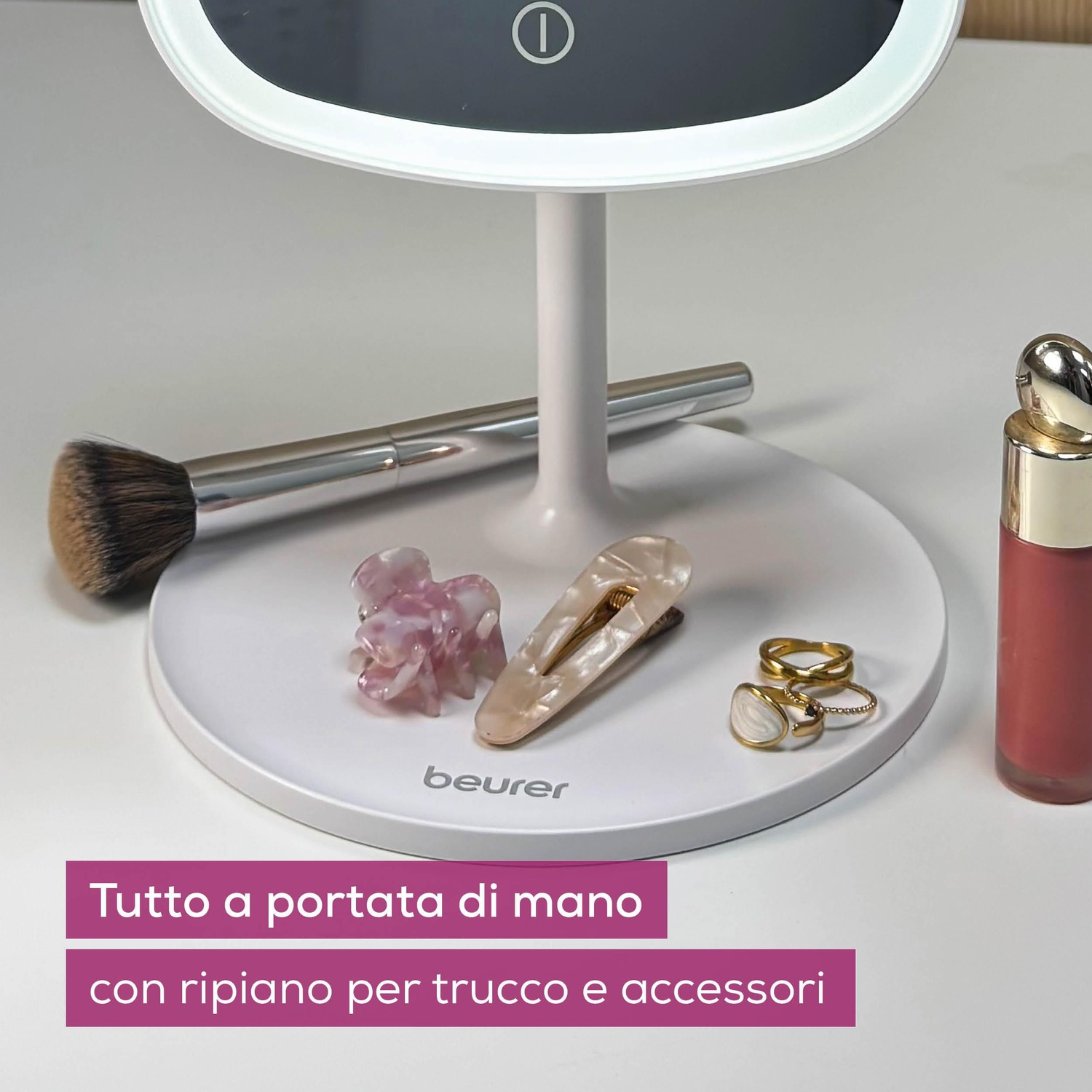 Uno specchio per il trucco bianco con accessori come pennelli, fermagli per capelli, anelli e rossetto su una superficie bianca.