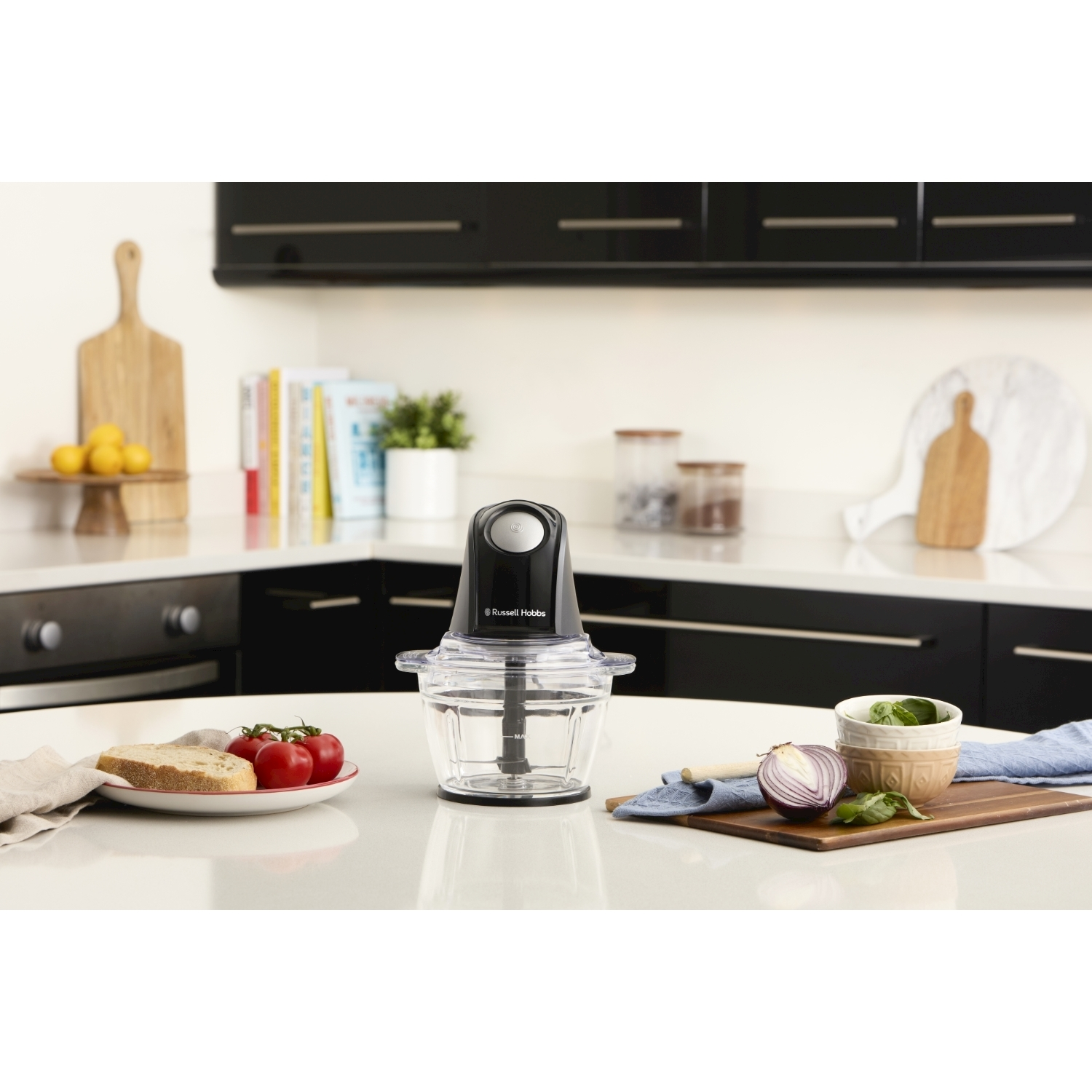 Scena di cucina con un robot da cucina, pomodori, pane e cipolla su un piano di lavoro bianco. Lo sfondo include mobili e mensole.