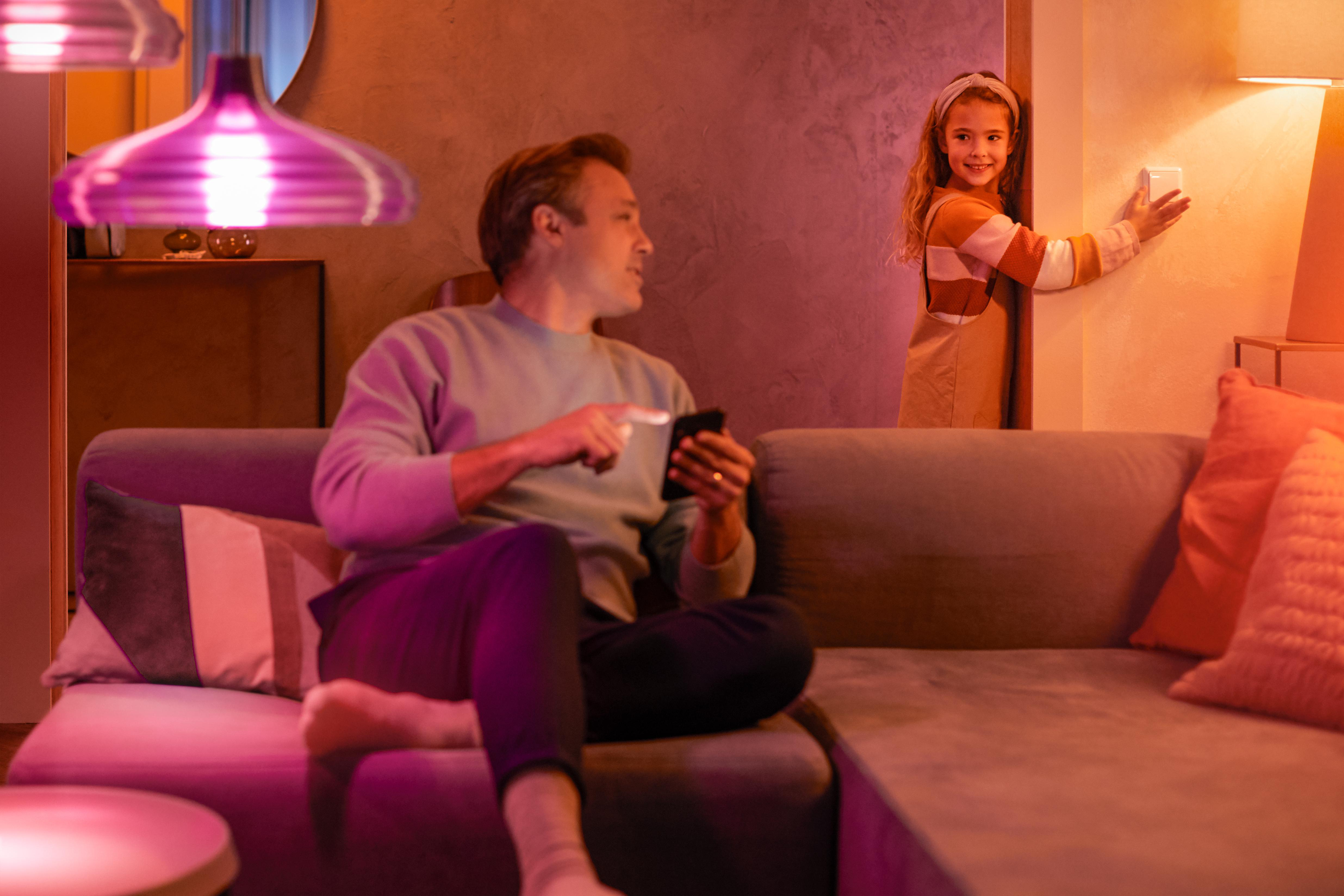 Mann auf Sofa mit Handy. Mädchen am Schalter. Rosa Licht von Lampen. Kissen und Wand im Hintergrund.