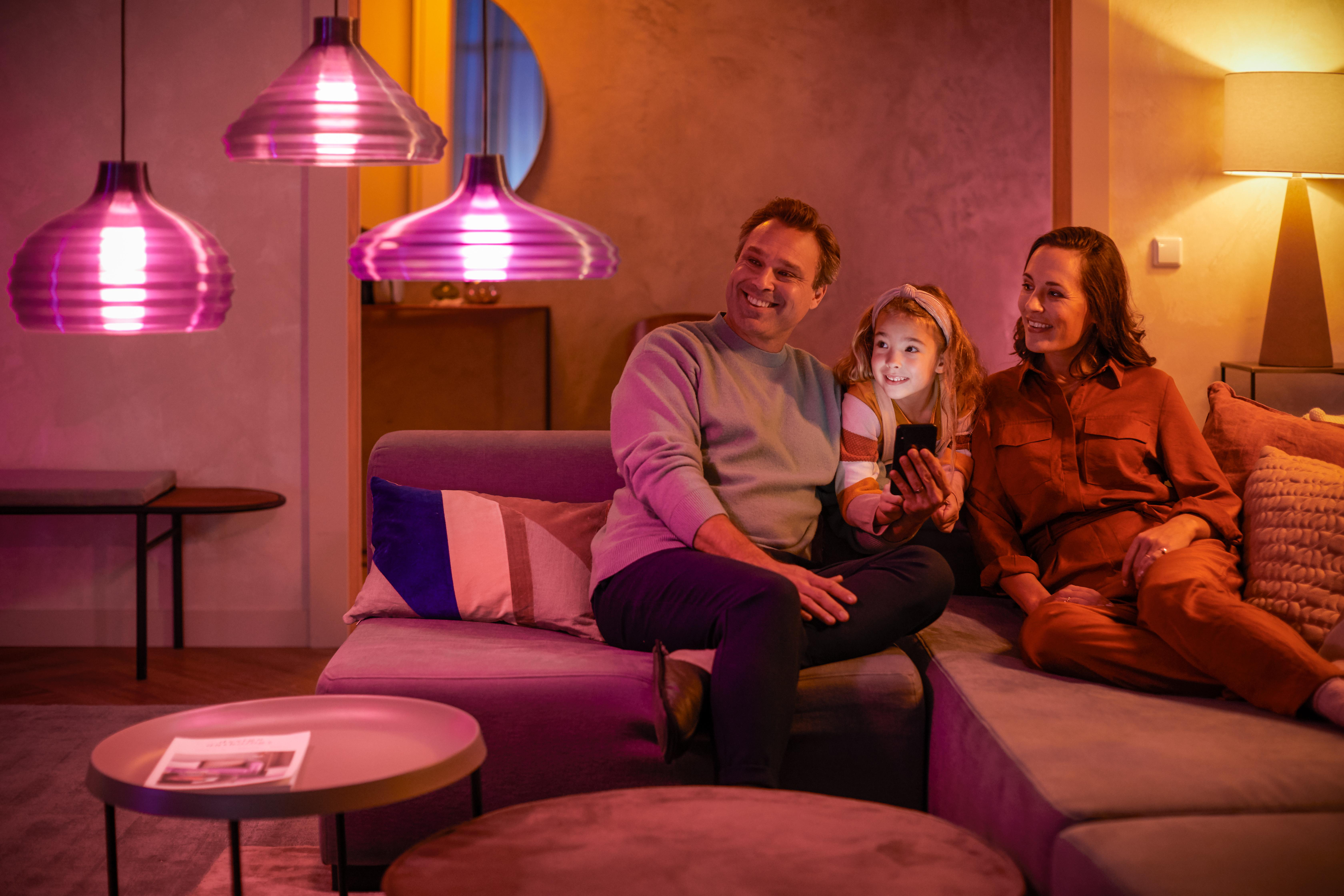 Familie sitzt auf einem Sofa und schaut auf ein Telefon. Rosa Lampen hängen darüber. Warme Beleuchtung.