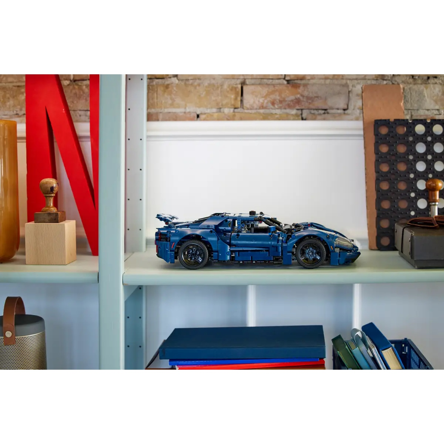 A blue Lego car sits on a shelf. Other items include books, a speaker, and decorative objects. The background features a brick wall.
