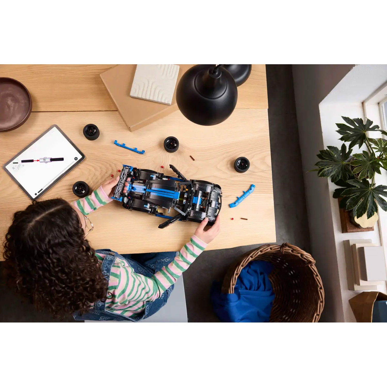 A person assembles a black and blue toy car on a wooden table. Wheels and parts are scattered around. A tablet and a plant are also visible.