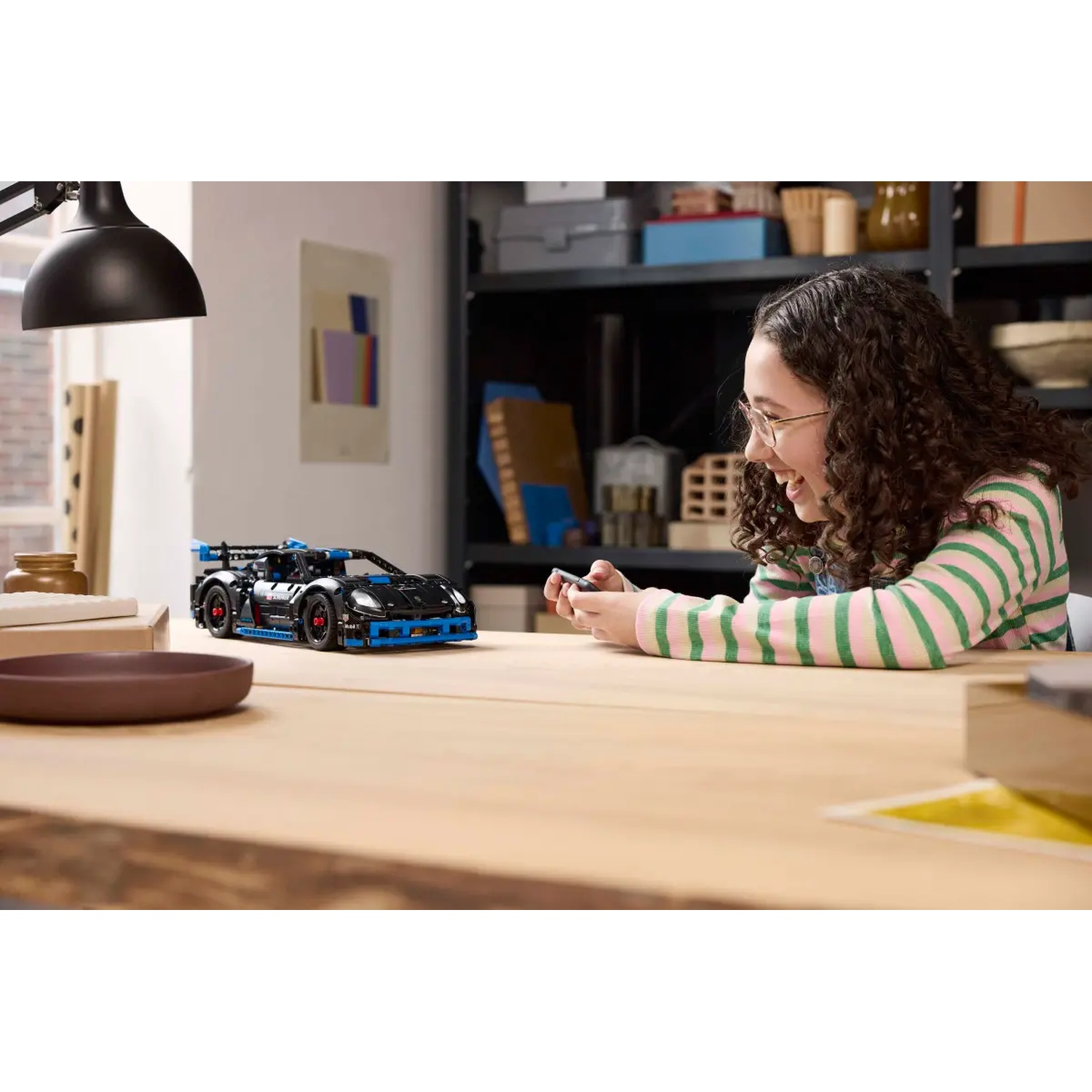 A girl with glasses smiles while holding a remote, controlling a black LEGO car on a wooden desk. A lamp, and shelves are in the background.