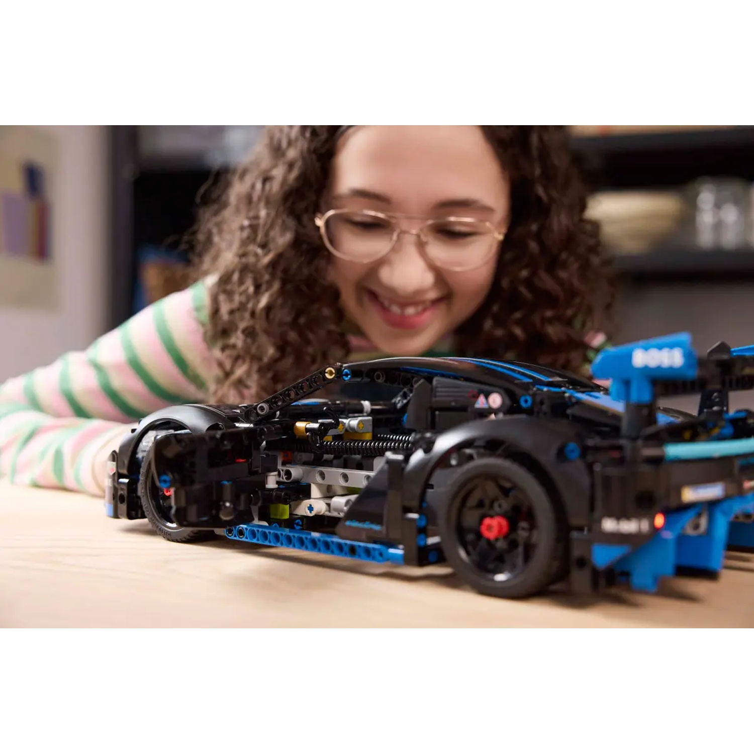 A girl smiles at a black and blue LEGO car on a wooden table. The car has visible mechanical components and red accents.
