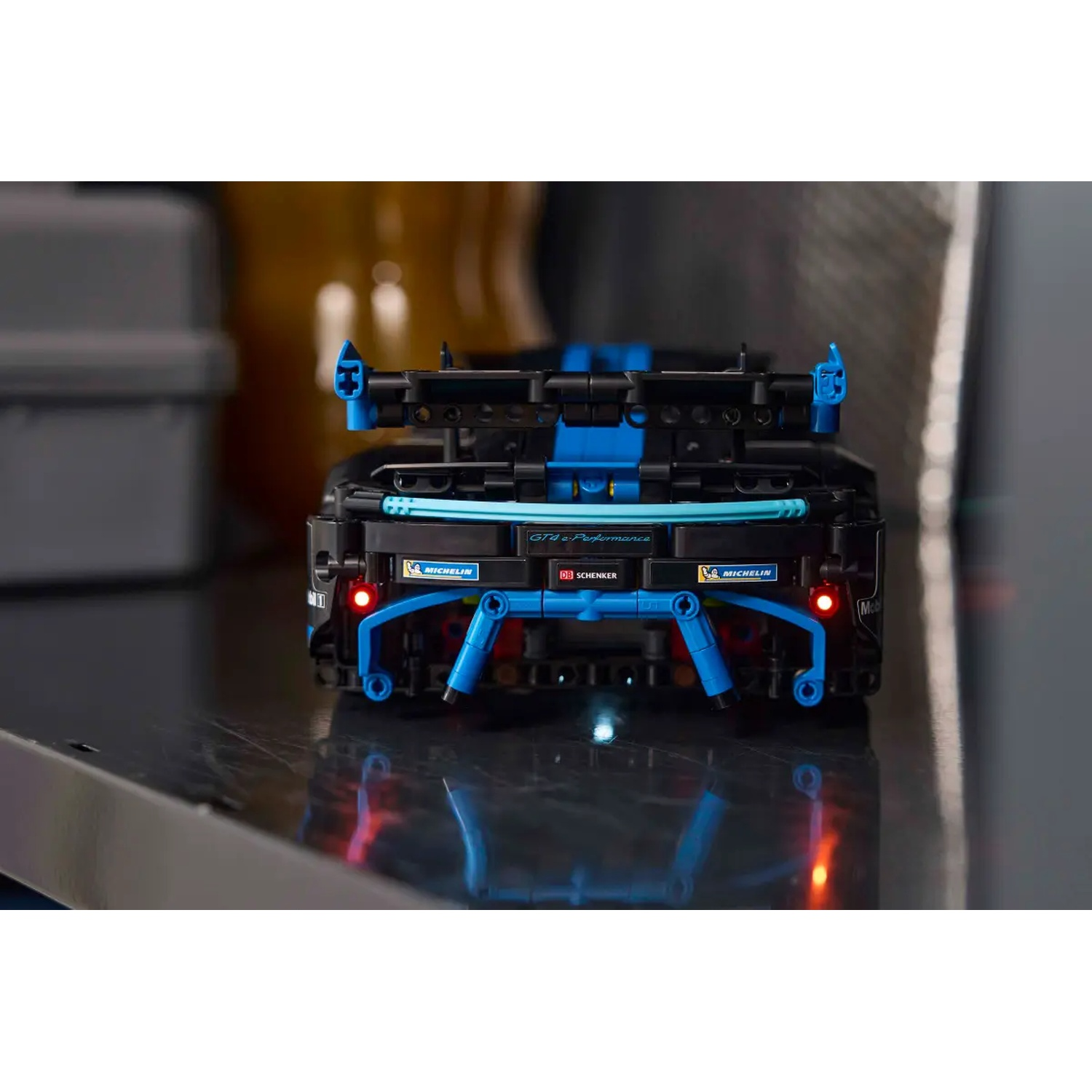 A close-up of a black and blue toy car. The rear of the car has blue accents and red lights. The car is on a reflective surface.