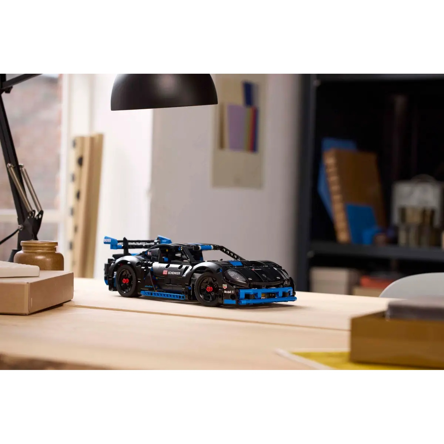 A black and blue Lego sports car sits on a wooden desk. A lamp and office supplies are in the background.