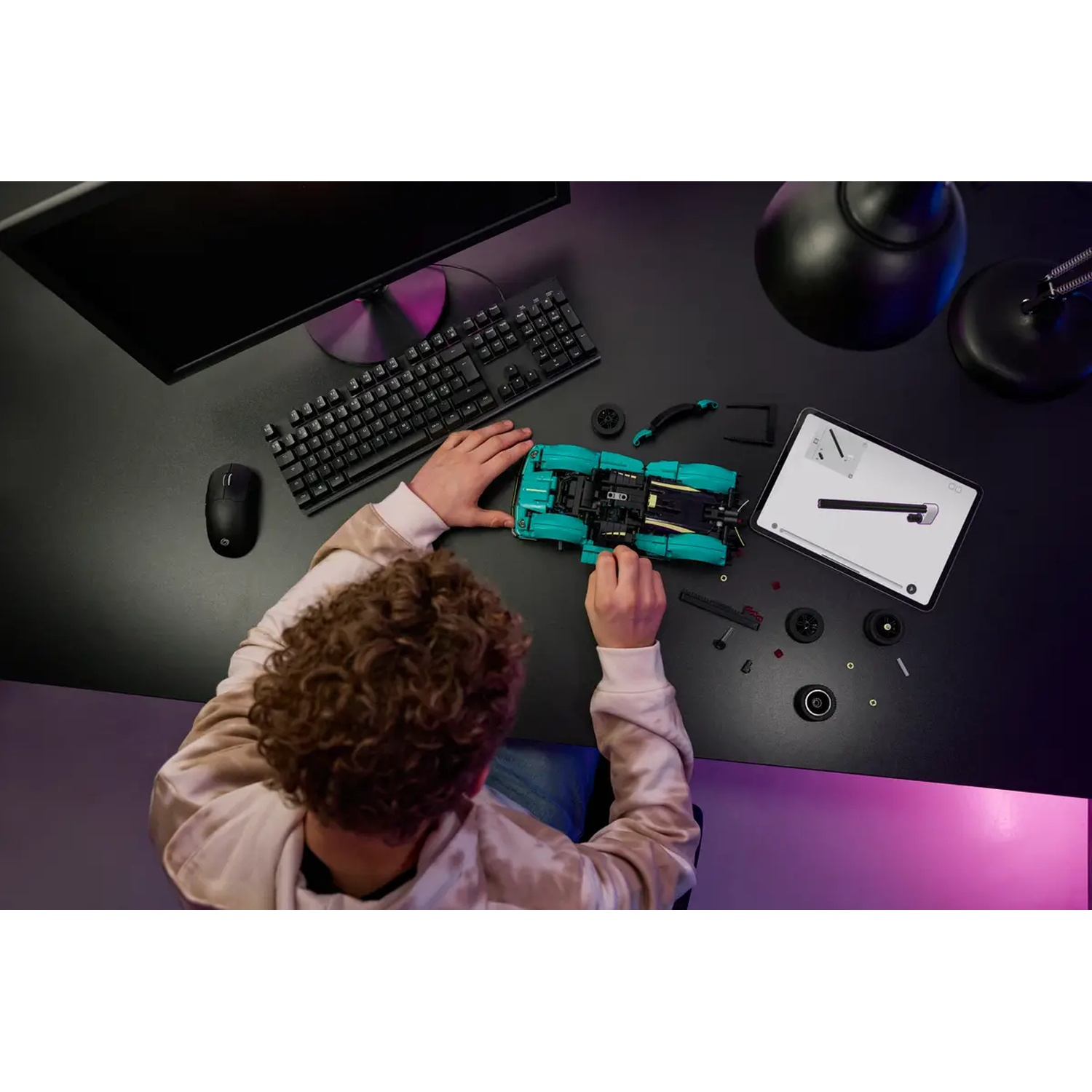 Een persoon monteert een turquoise Lego-auto op een zwart bureau. Er zijn ook een monitor, toetsenbord, muis en tablet aanwezig, met een lamp.