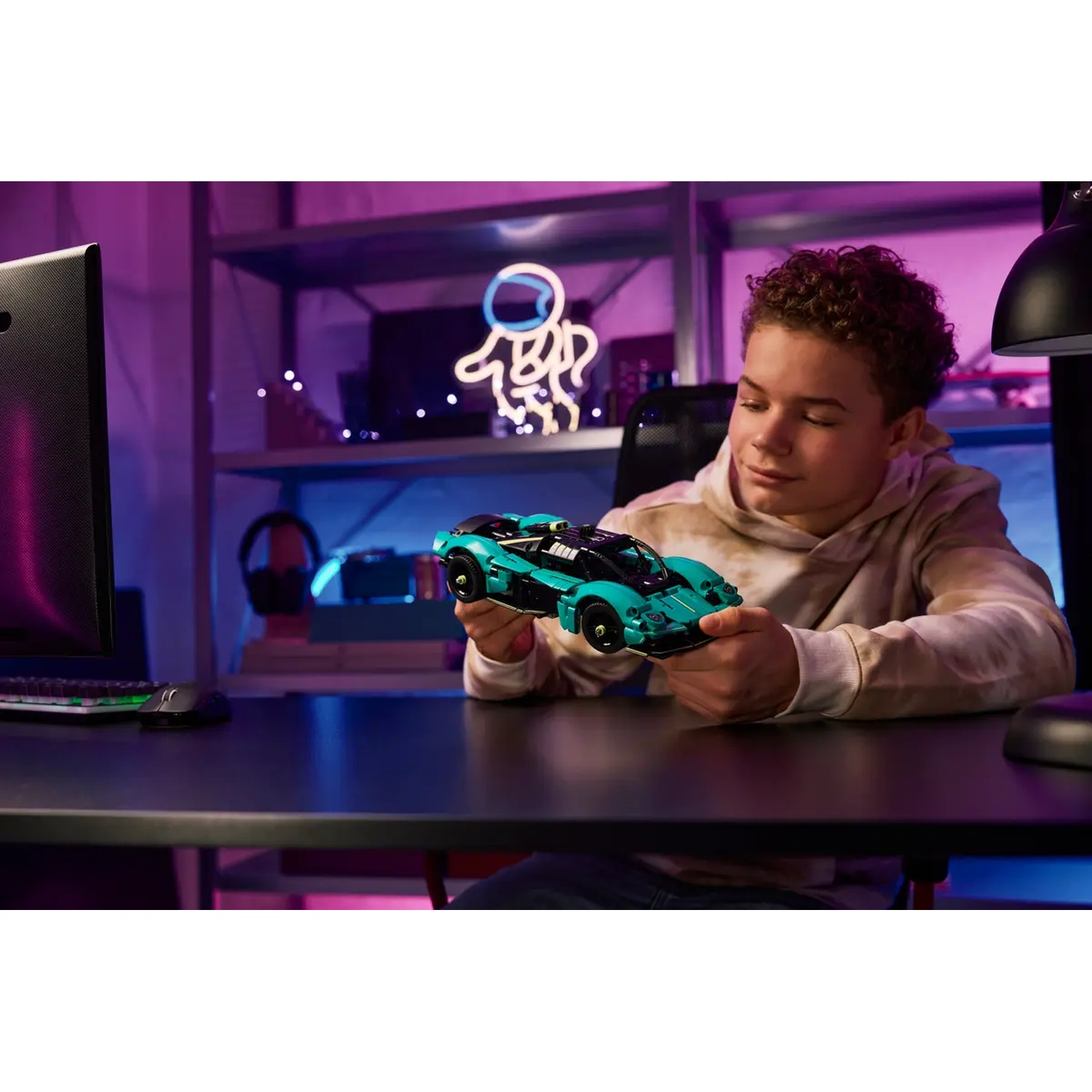Een jongen houdt een teal en zwarte speelgoedauto vast. Hij zit aan een bureau met een computer en een muis. Achtergrond bevat planken en een neonreclame.