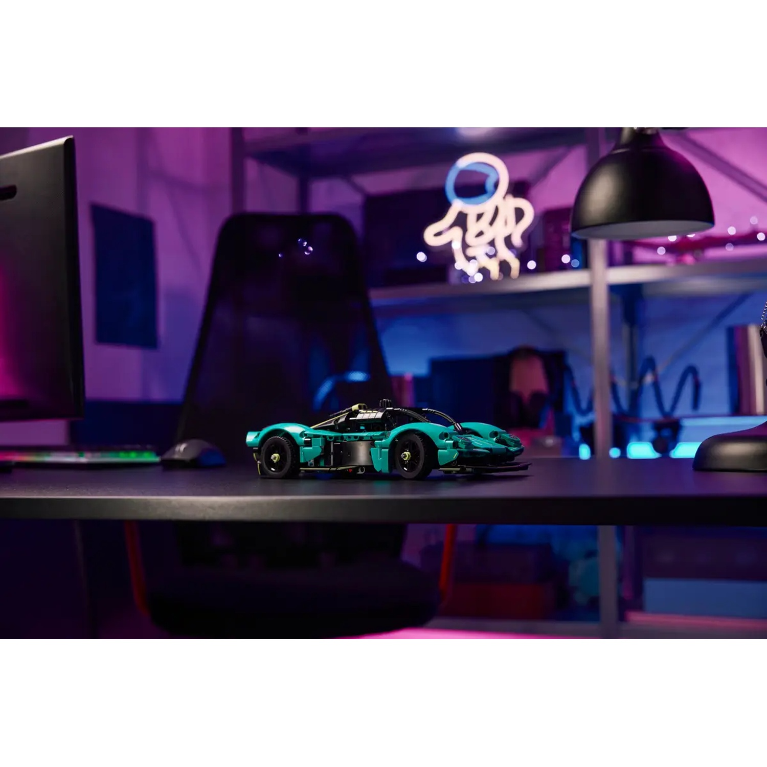 Een Lego sportwagen staat op een bureau. De scène omvat een computer, een stoel, een neon astronautenbord en een lamp.