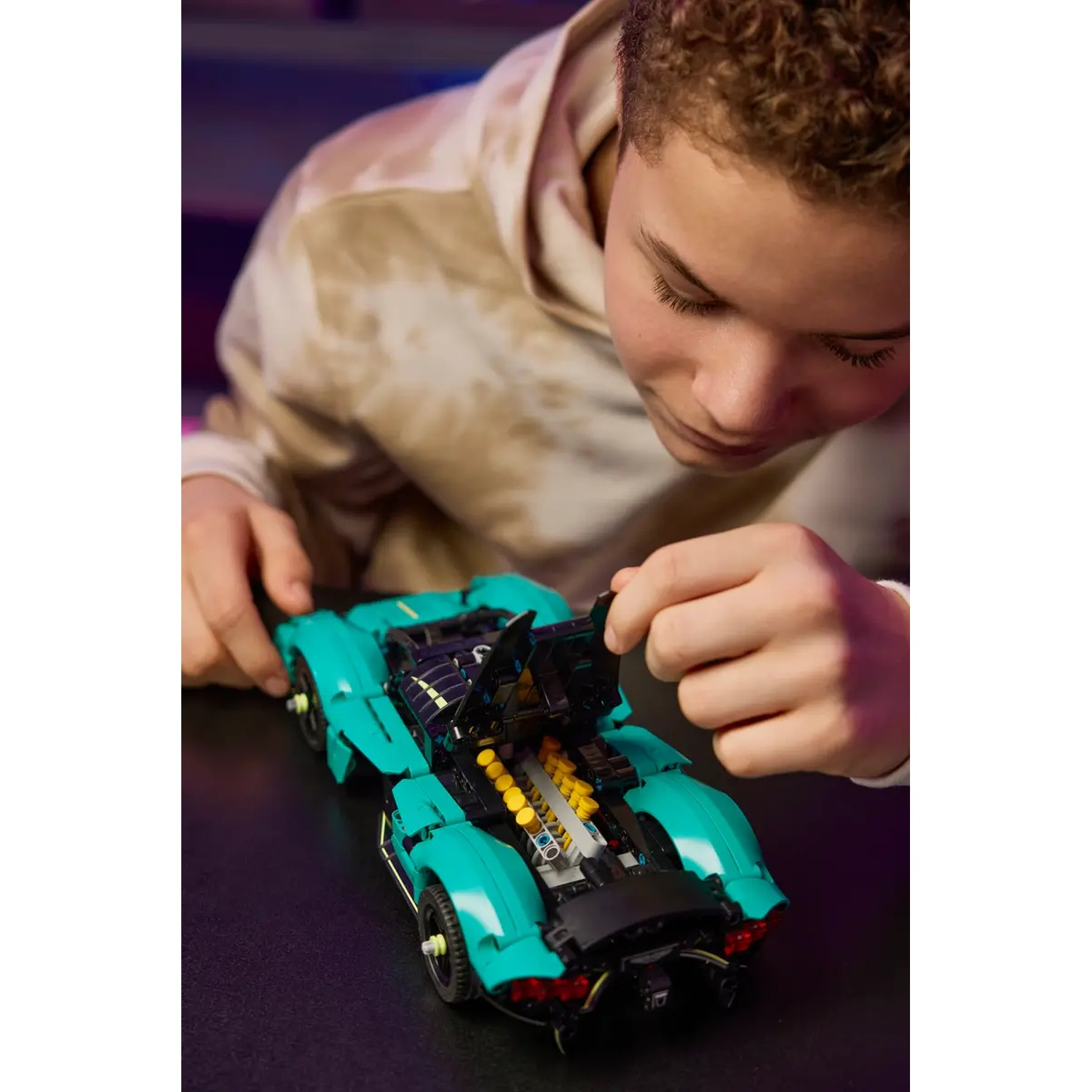 Een persoon werkt aan een turquoise speelgoedauto. De motorkap van de auto staat open. De persoon draagt een lichtbruine hoodie.