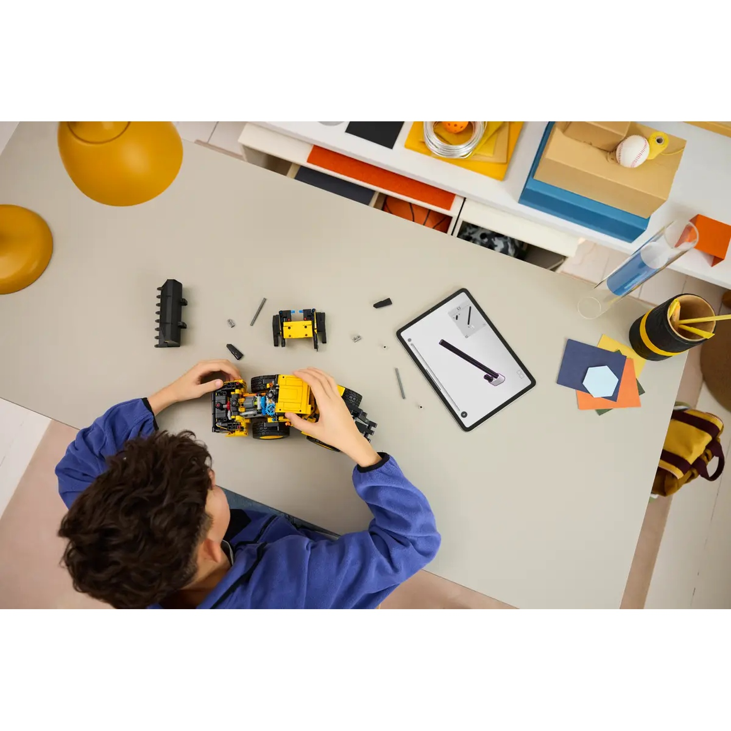 A child is building a yellow construction toy on a desk. Other toys and a tablet are visible. The scene is top-down.