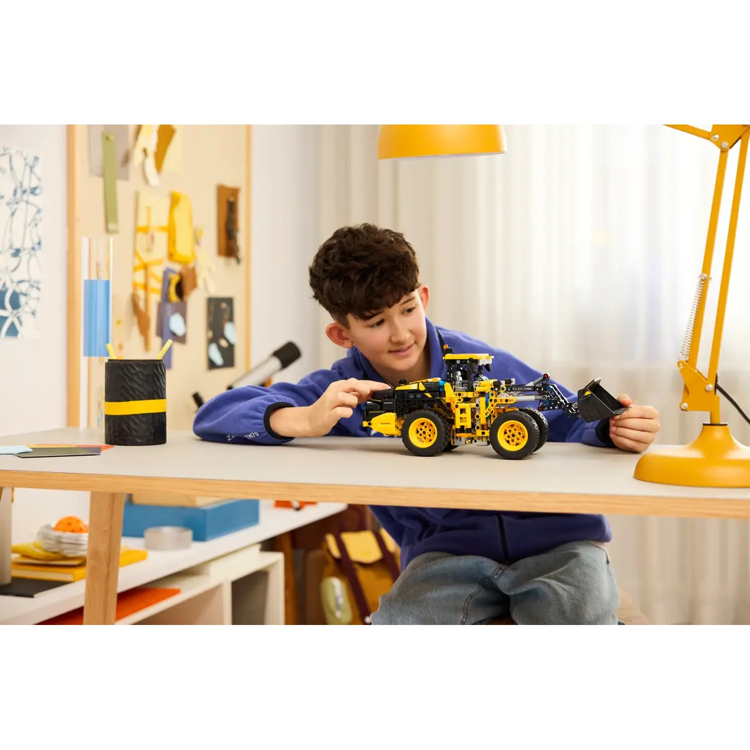 A boy in blue shirt plays with a yellow and black toy truck on a table. A yellow lamp and desk items are visible.