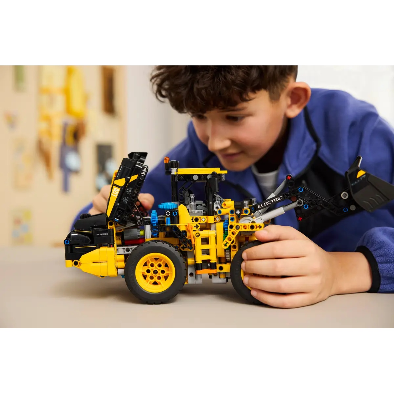 A boy is focused on a yellow and black construction toy. The toy resembles a front loader. The boy is wearing a blue jacket.