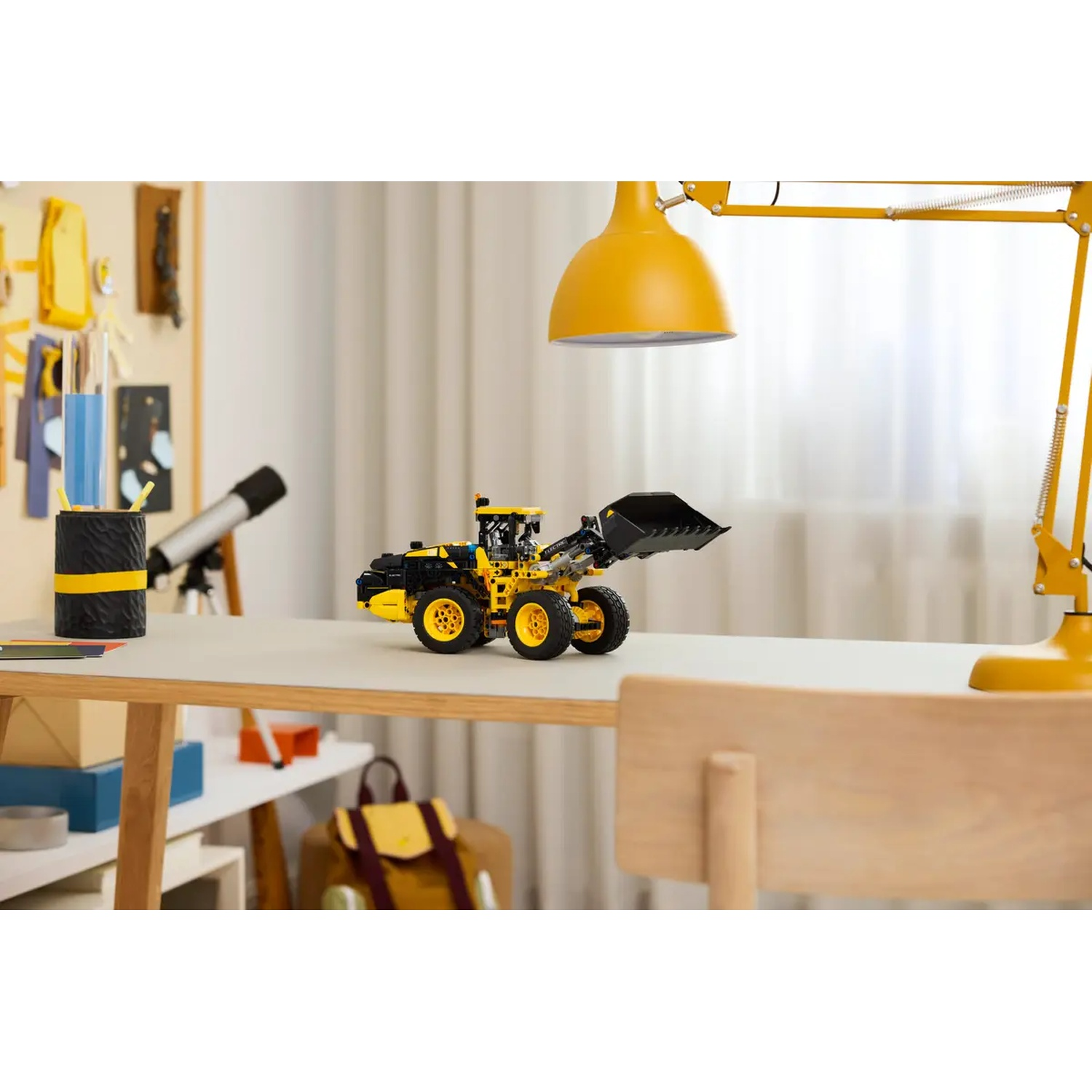 A yellow and black toy front loader sits on a white desk. A yellow lamp hangs above it. A chair and backpack are nearby.