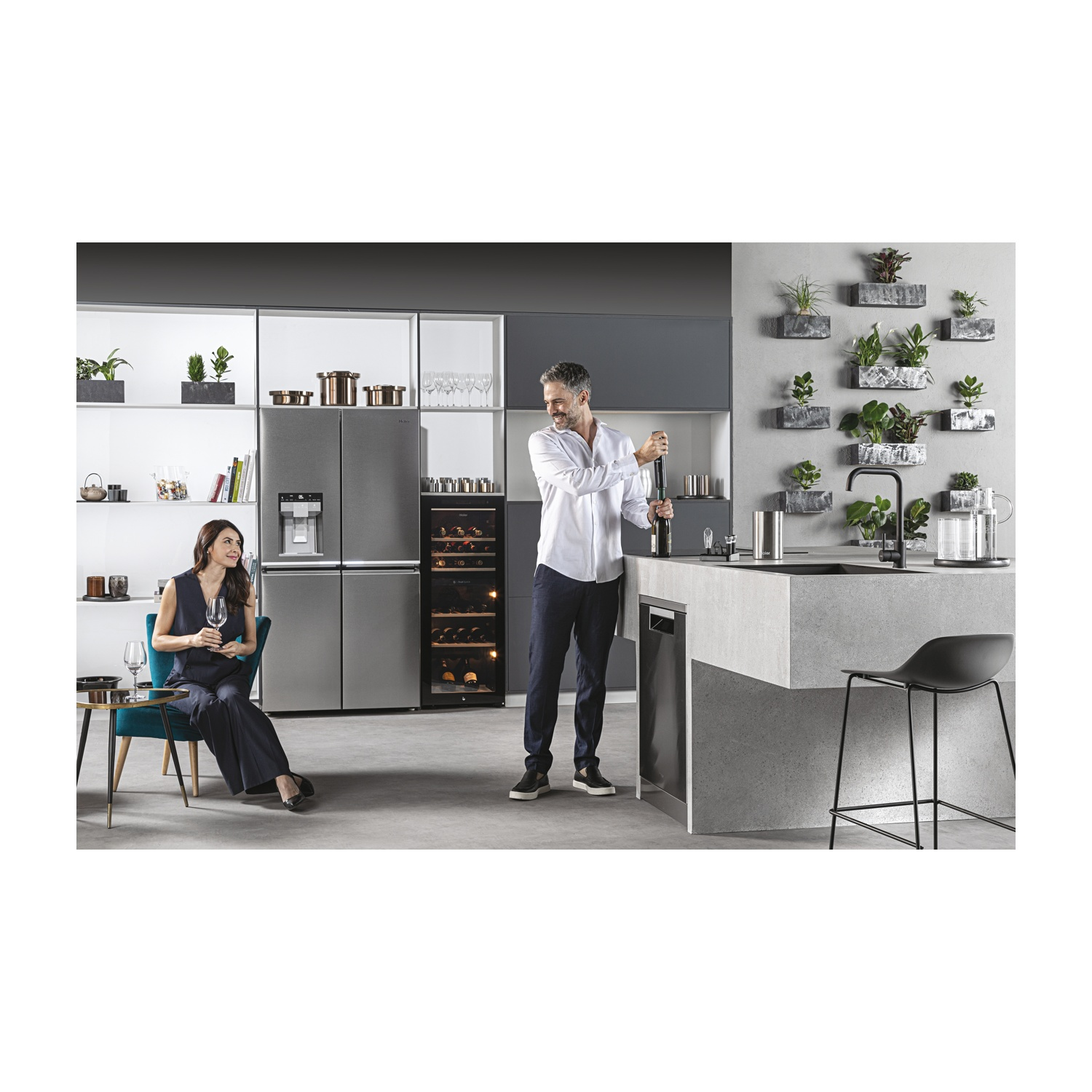 Un uomo apre una bottiglia di vino in una cucina moderna. Una donna è seduta, con un bicchiere in mano. Le piante adornano il muro. Elettrodomestici in acciaio inox.