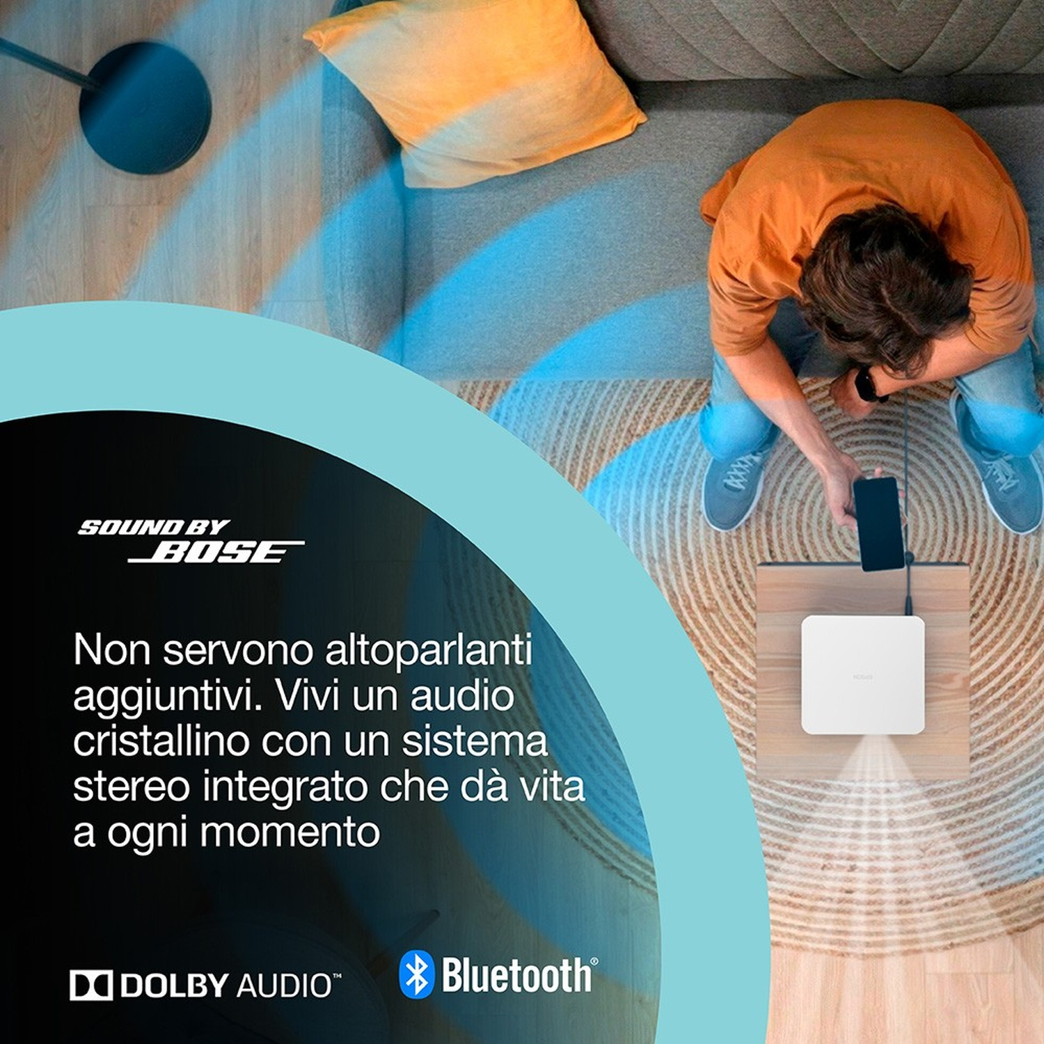 Vista dall'alto: Uomo con telefono si collega a altoparlante bianco. Divano con cuscino giallo visibile.