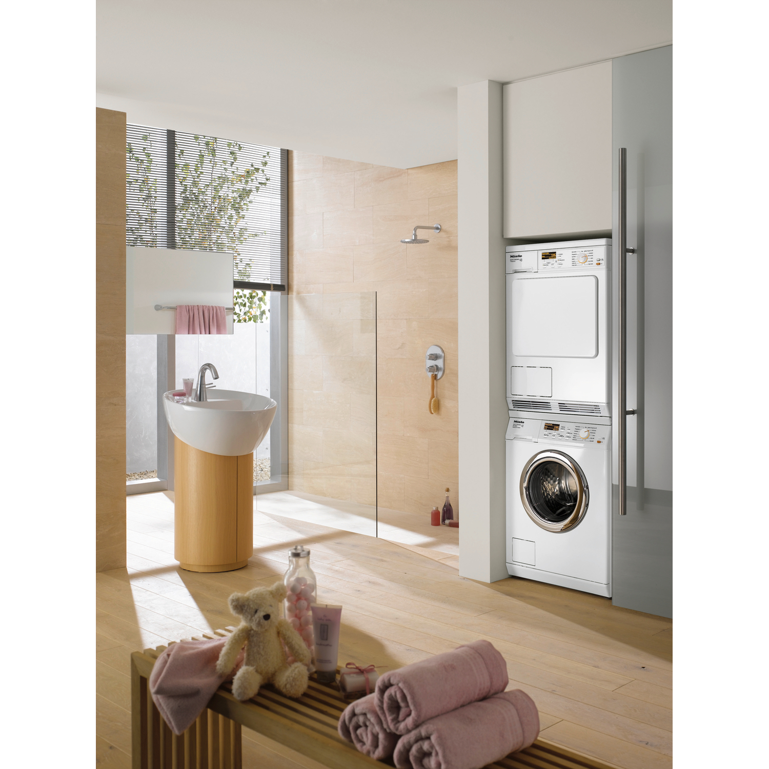 Badkamer met wasmachine, droger, wastafel, handdoeken, een teddybeer en een houten bank. Lichte bruintinten domineren.