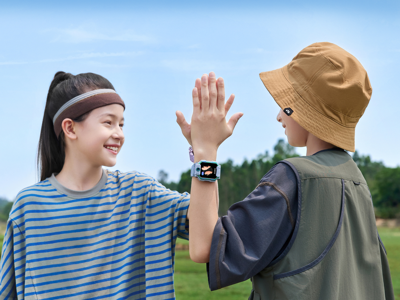 Zwei Kinder geben sich High-Five. Einer trägt ein gestreiftes Hemd, der andere einen Hut. Beide haben Smartwatches. Blauer Himmel und grüne Bäume im Hintergrund.