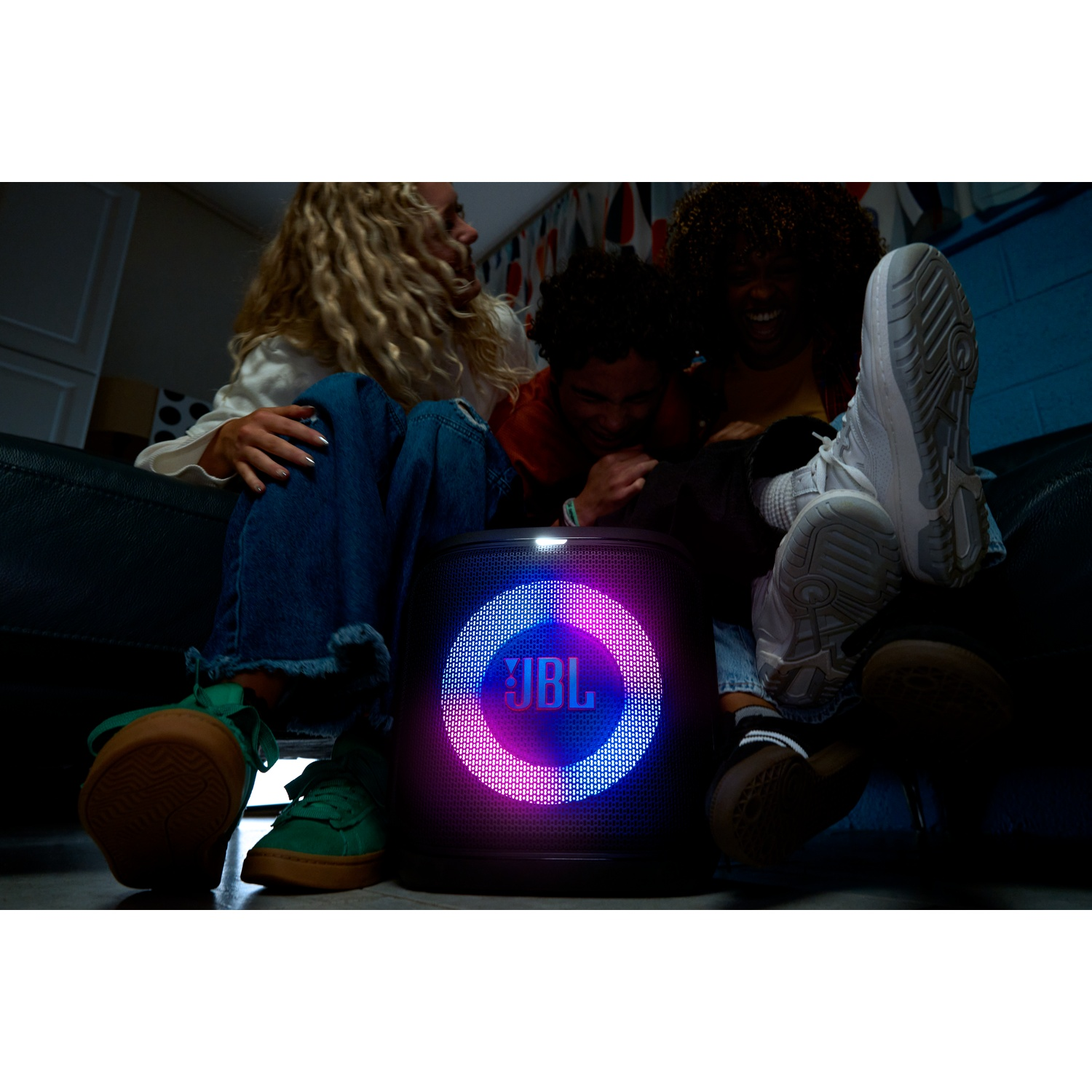 Three people seated around a glowing JBL speaker. The speaker emits pink and blue light. People are laughing.