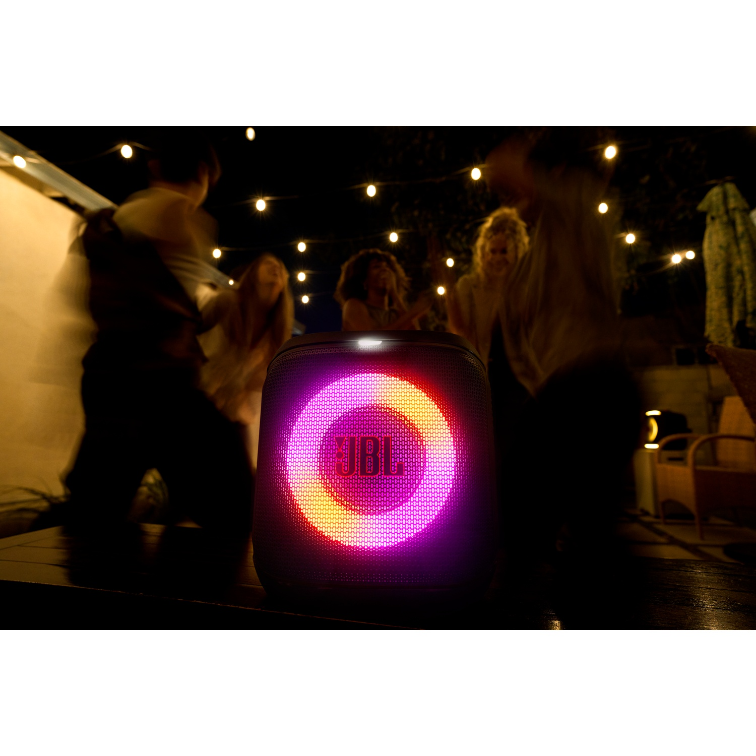 A JBL speaker glows with colorful lights as blurred figures dance nearby under string lights at night.